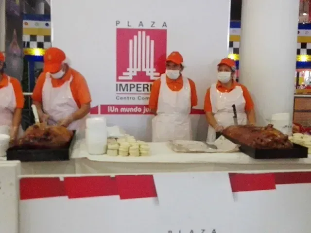 Tres cocineros con uniformes naranjas sirven carne asada en un mostrador de bufé, con un letrero de Plaza Imperial detrás de ellos