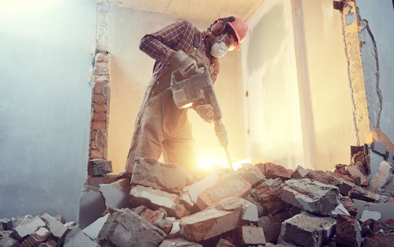 Worker demolishing a wall amid rubble in a bright, damaged room