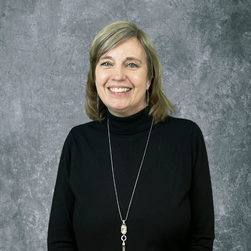 Smiling woman in a black turtleneck and long necklace against a gray backdrop