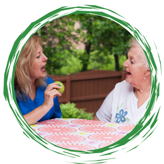Two people talking at a table outdoors, with a green garden and wooden fence behind them.