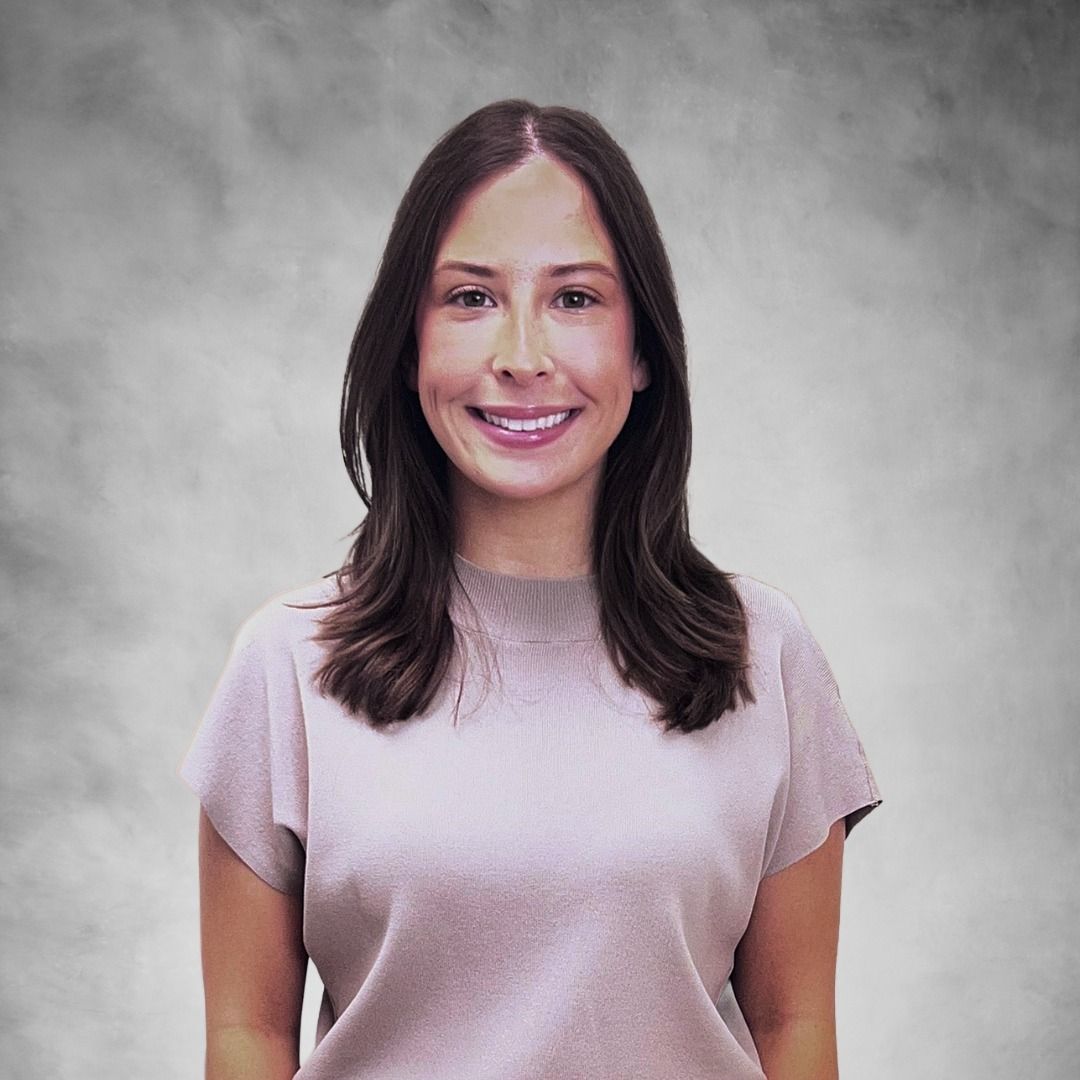 Smiling person with long dark hair wearing a light pink shirt against a gray studio backdrop