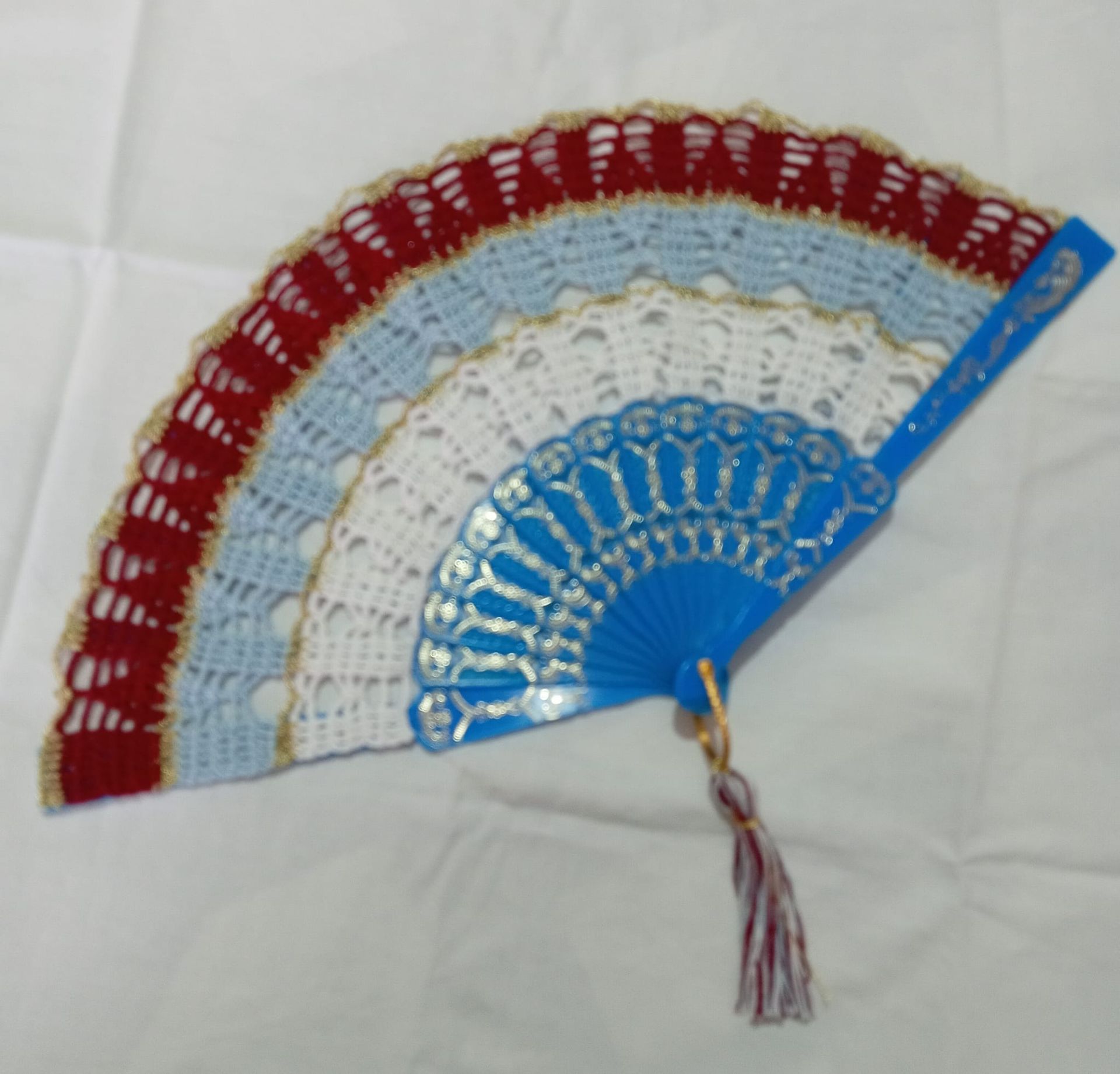 Ventaglio pieghevole decorativo con pannelli decorati nei toni del blu, bianco e rosso e nappa su sfondo bianco.