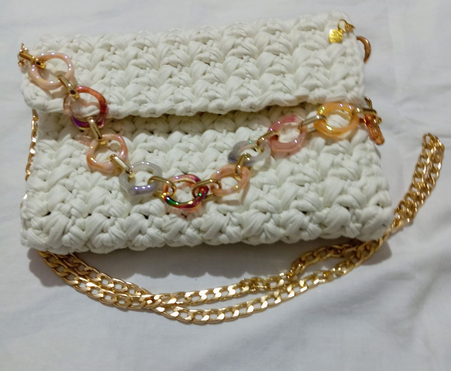 Pochette bianca all'uncinetto con catena dorata e collana a maglie pastello su sfondo chiaro