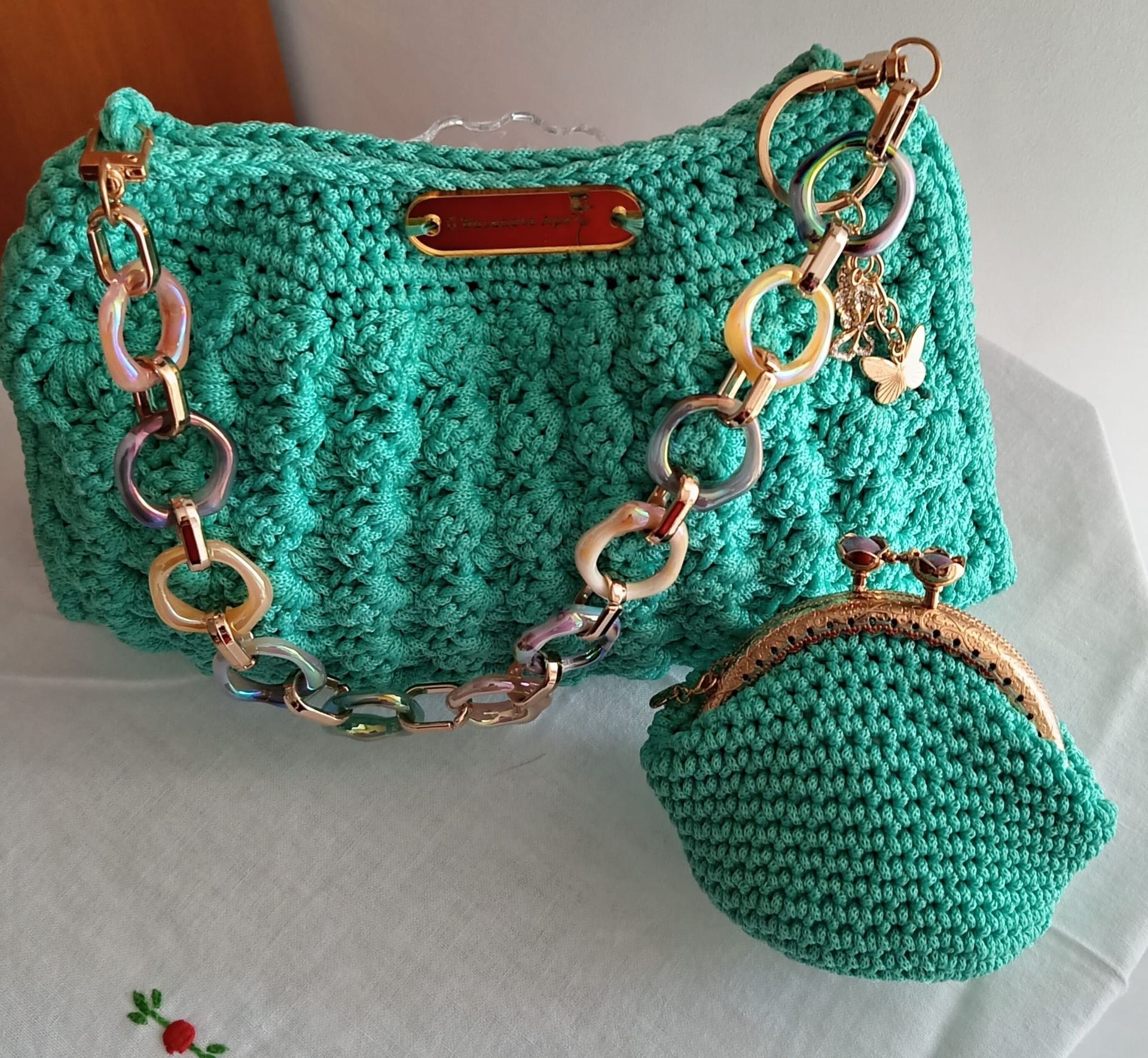 Borsa a mano turchese all'uncinetto con catena e chiusura dorate, e portamonete abbinato su sfondo chiaro.