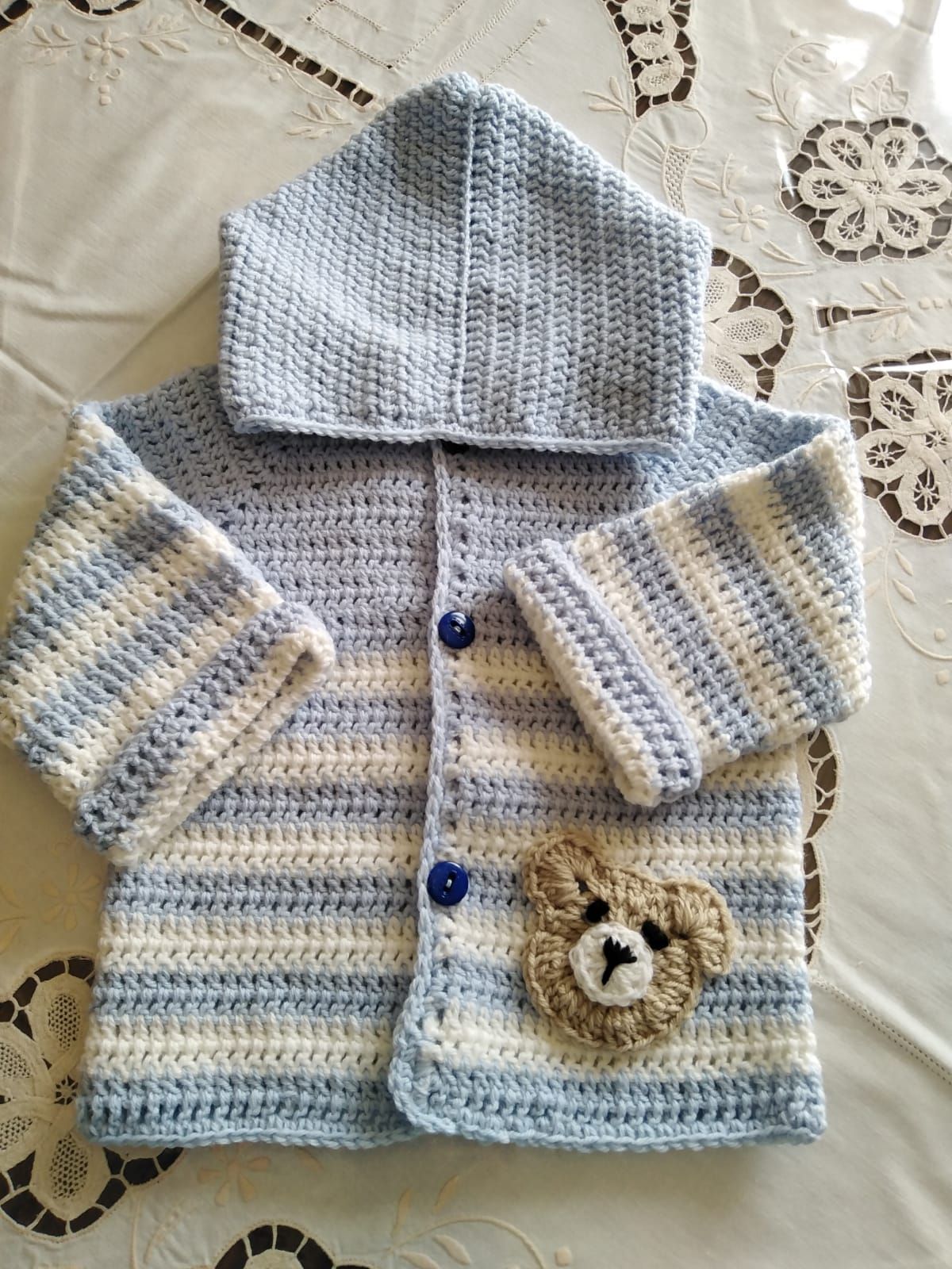 Felpa con cappuccio per neonati realizzata a mano all'uncinetto, nei colori bianco, blu e giallo, con una piccola toppa a forma di orsetto su una tovaglia di pizzo.