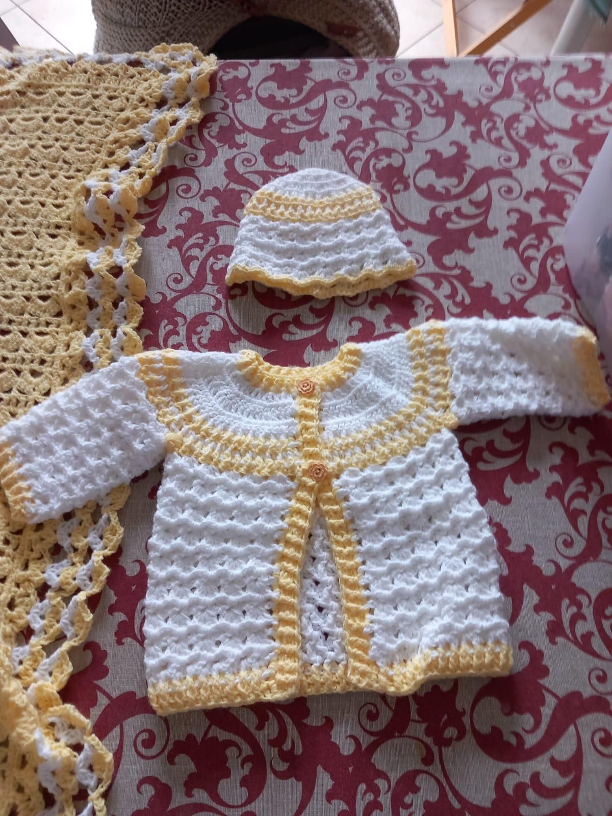 Maglioncino per neonato bianco e giallo lavorato all'uncinetto con cappellino abbinato su sfondo di tessuto fantasia.