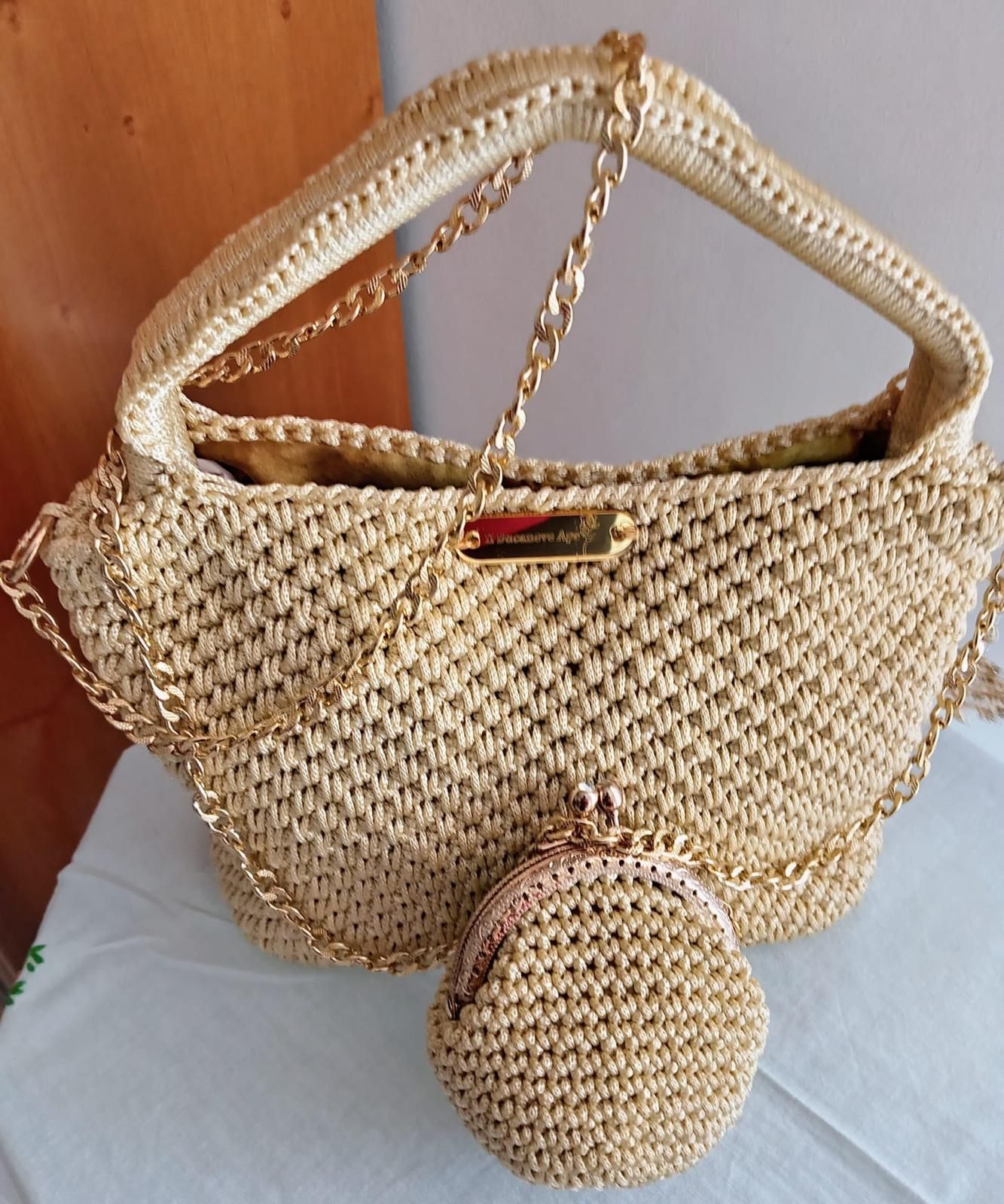 Borsa a mano beige intrecciata con manico a catena dorata e pochette rotonda abbinata su una base bianca.