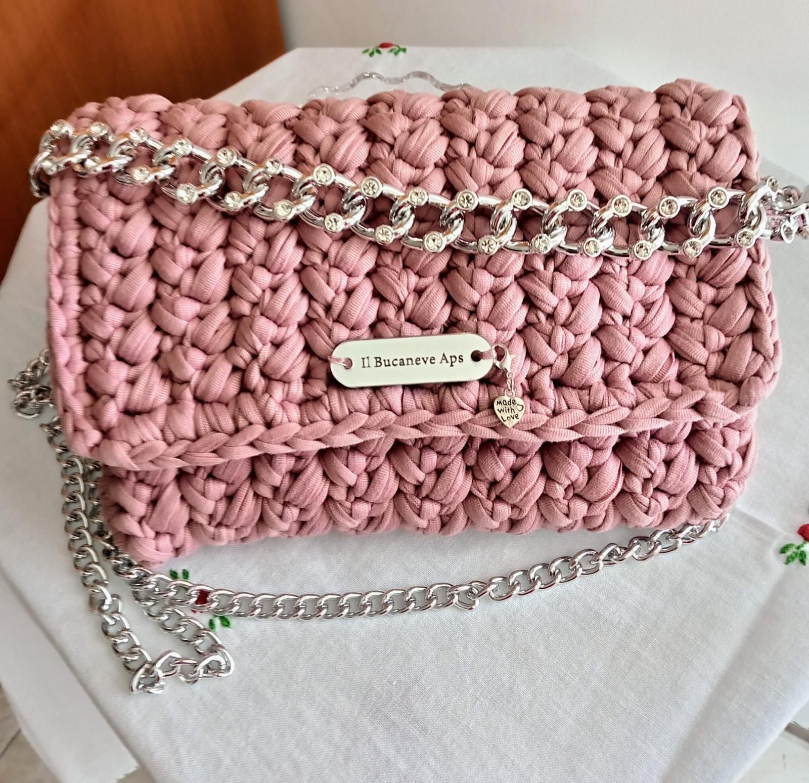 Borsa rosa intrecciata con tracolla a catena argentata e chiusura a moschettone su tessuto bianco.