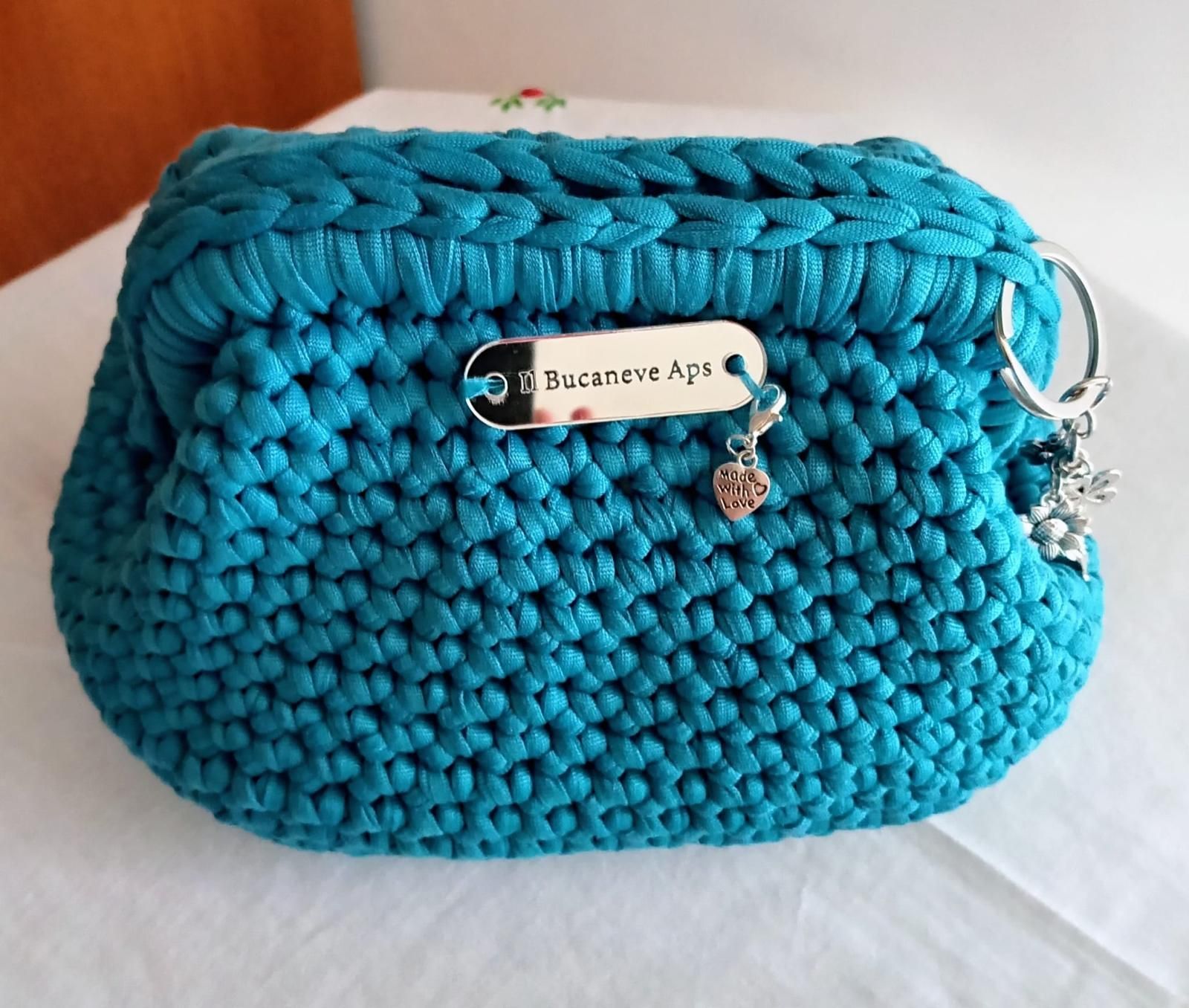 Borsa all'uncinetto turchese con targhetta e ciondolo argentati su sfondo bianco.