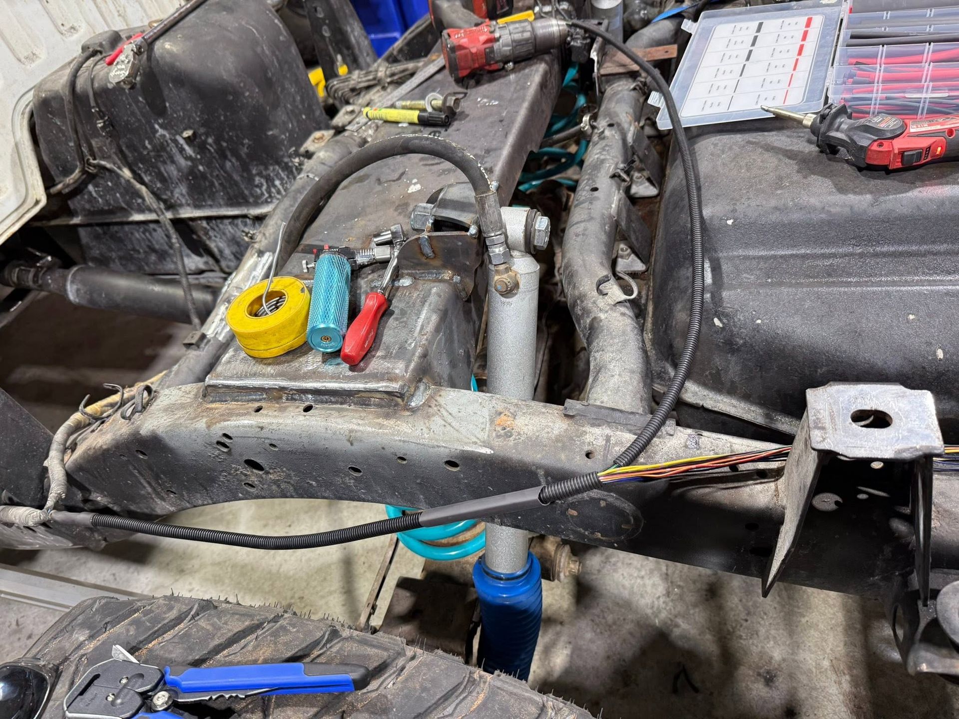 Close-up of a vehicle engine bay with tools, wiring, and a yellow tape measure on a dirty metal frame.
