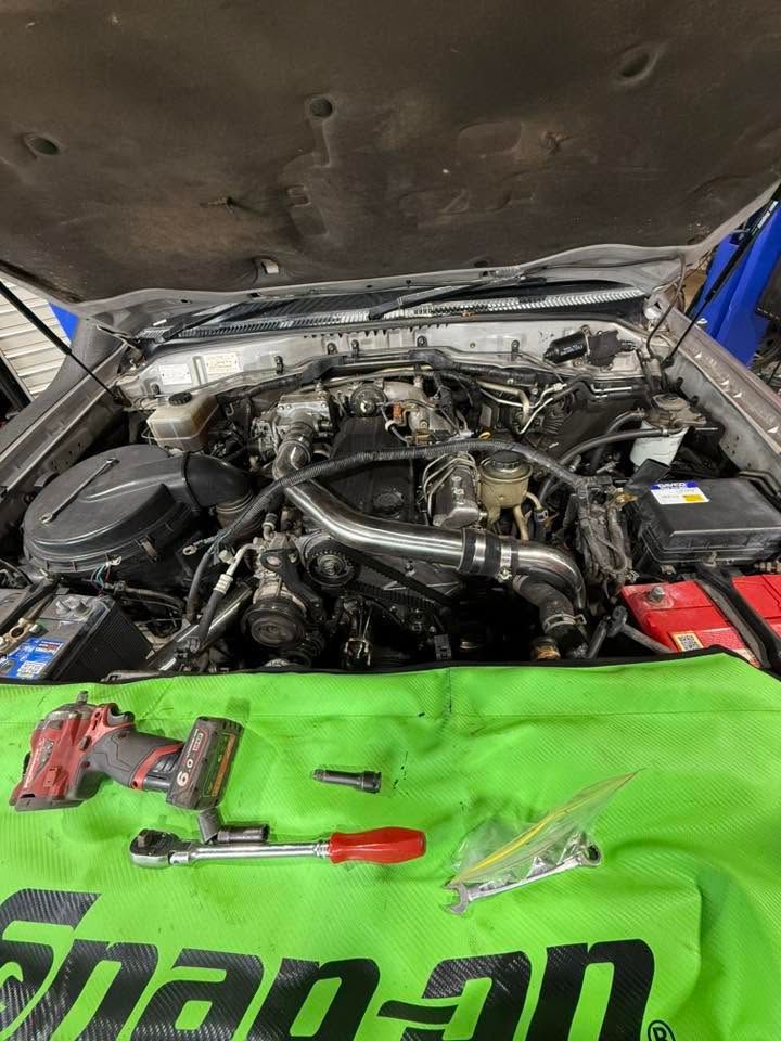 Car engine bay with hood open, tools on a green fender cover, and red battery visible on the right.