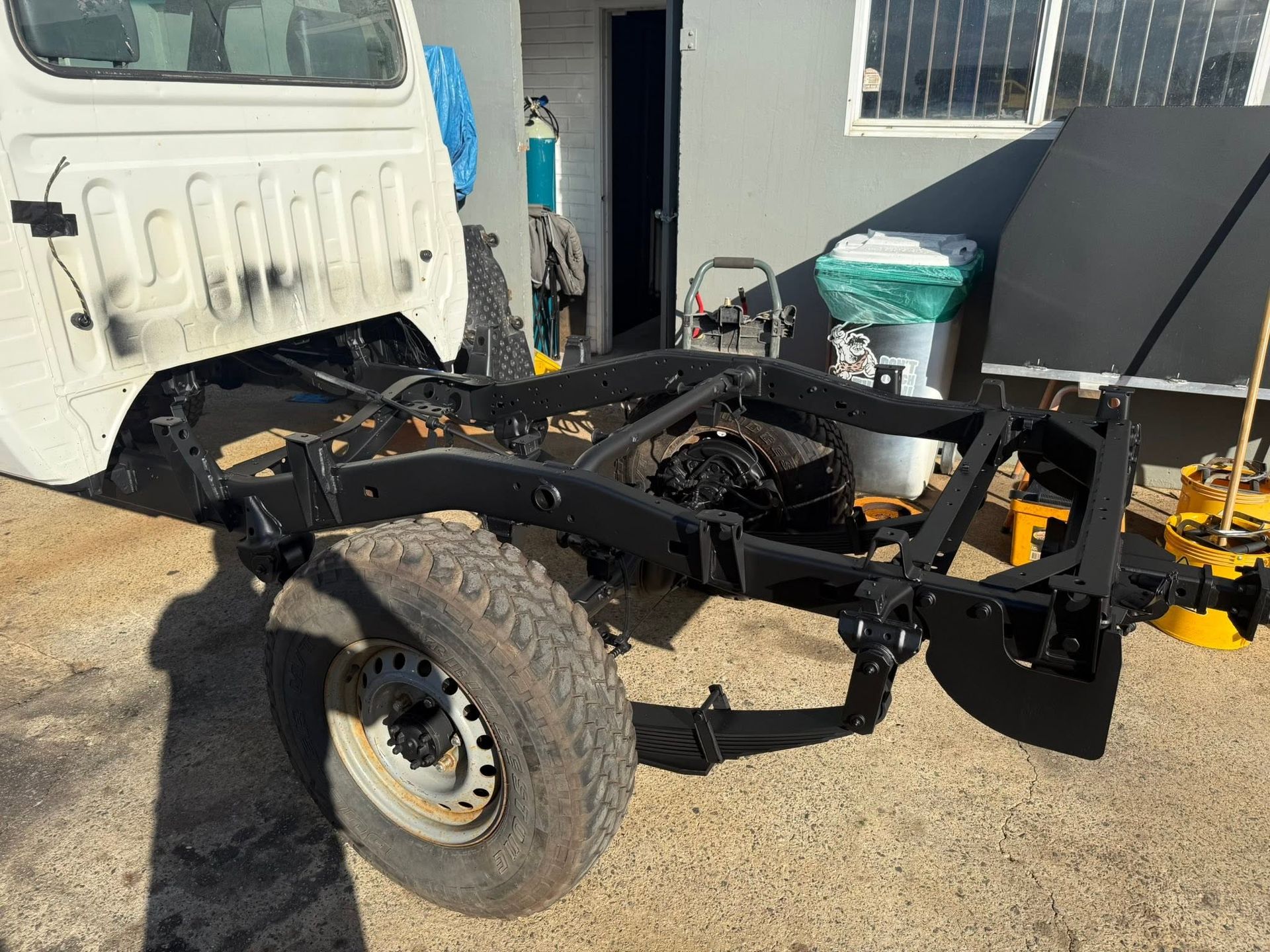 Bare truck chassis with large off-road tires parked beside a white building and gravel driveway