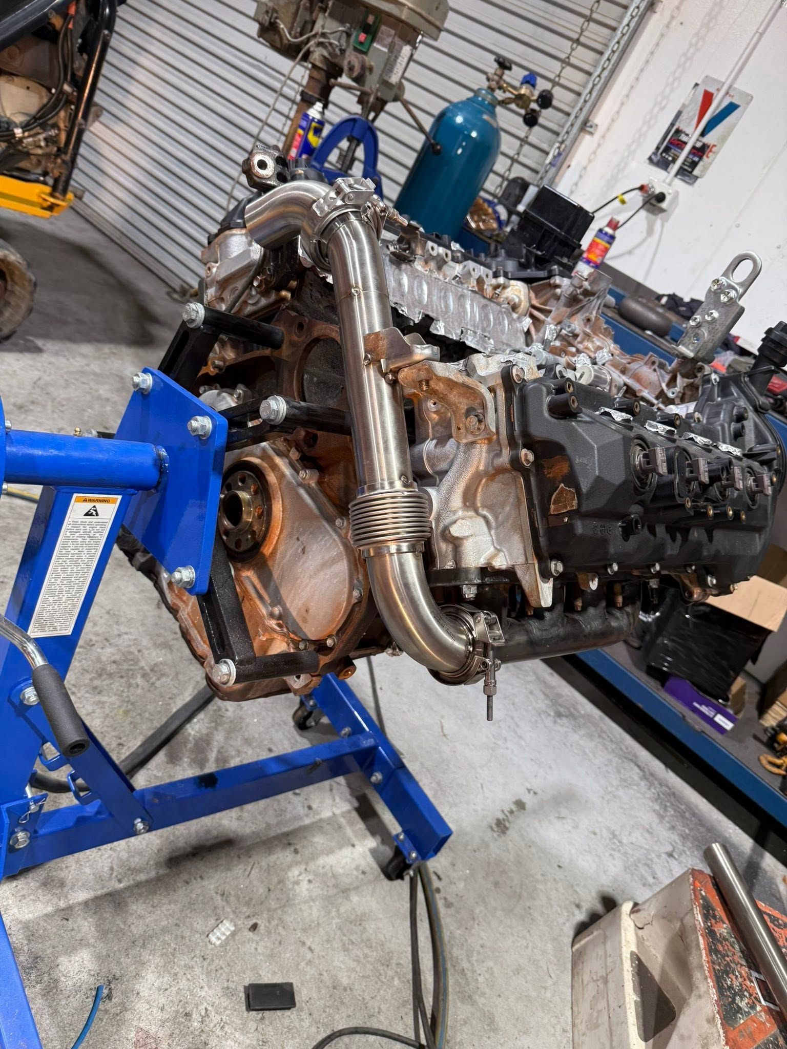 Engine on a blue stand in a workshop, with polished exhaust pipes and tools nearby.