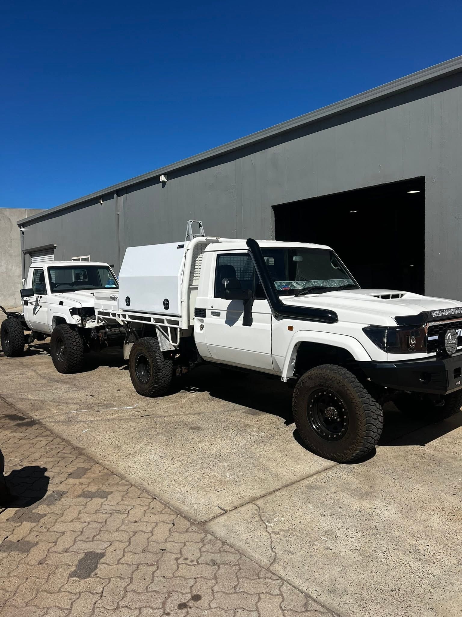 Two white utility trucks parked outside a warehouse on a sunny day