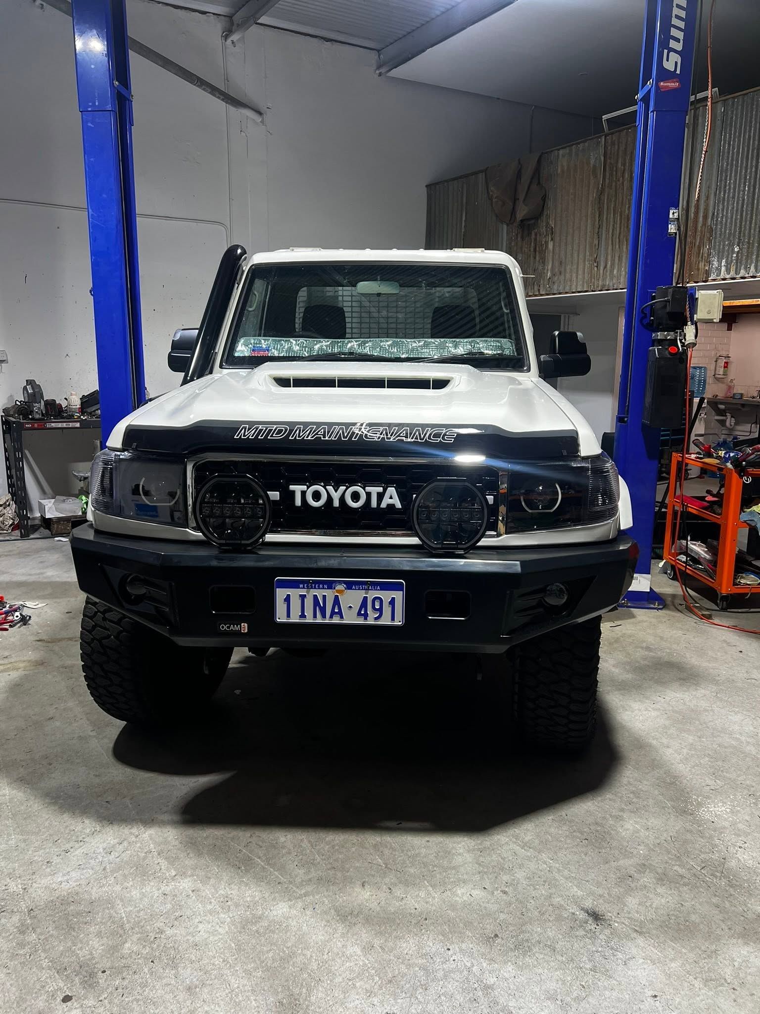White Toyota truck on a lift in a garage, viewed from the front with blue columns and tools nearby