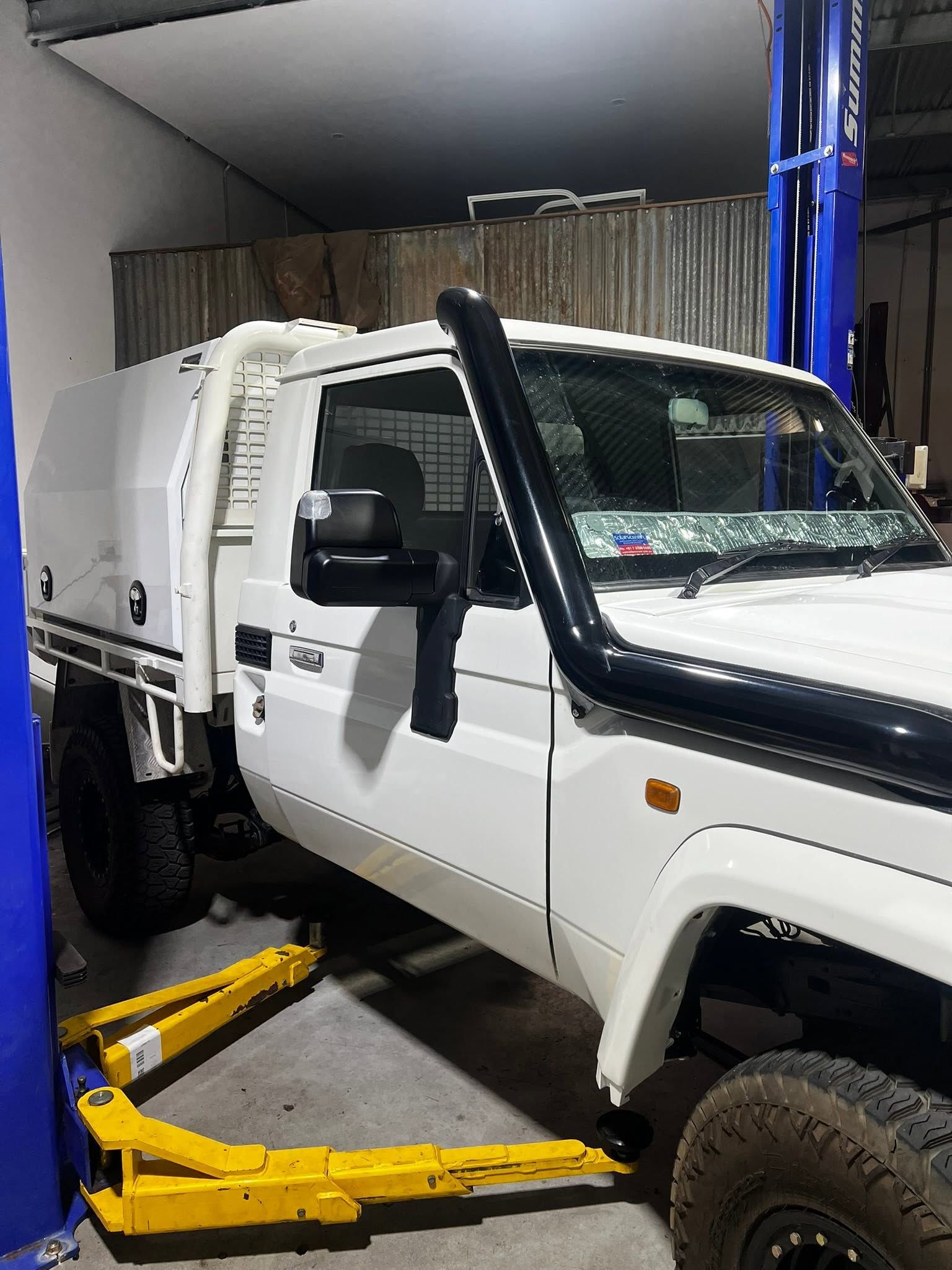White off-road utility truck on a lift in a garage, with a black snorkel and open driver door.