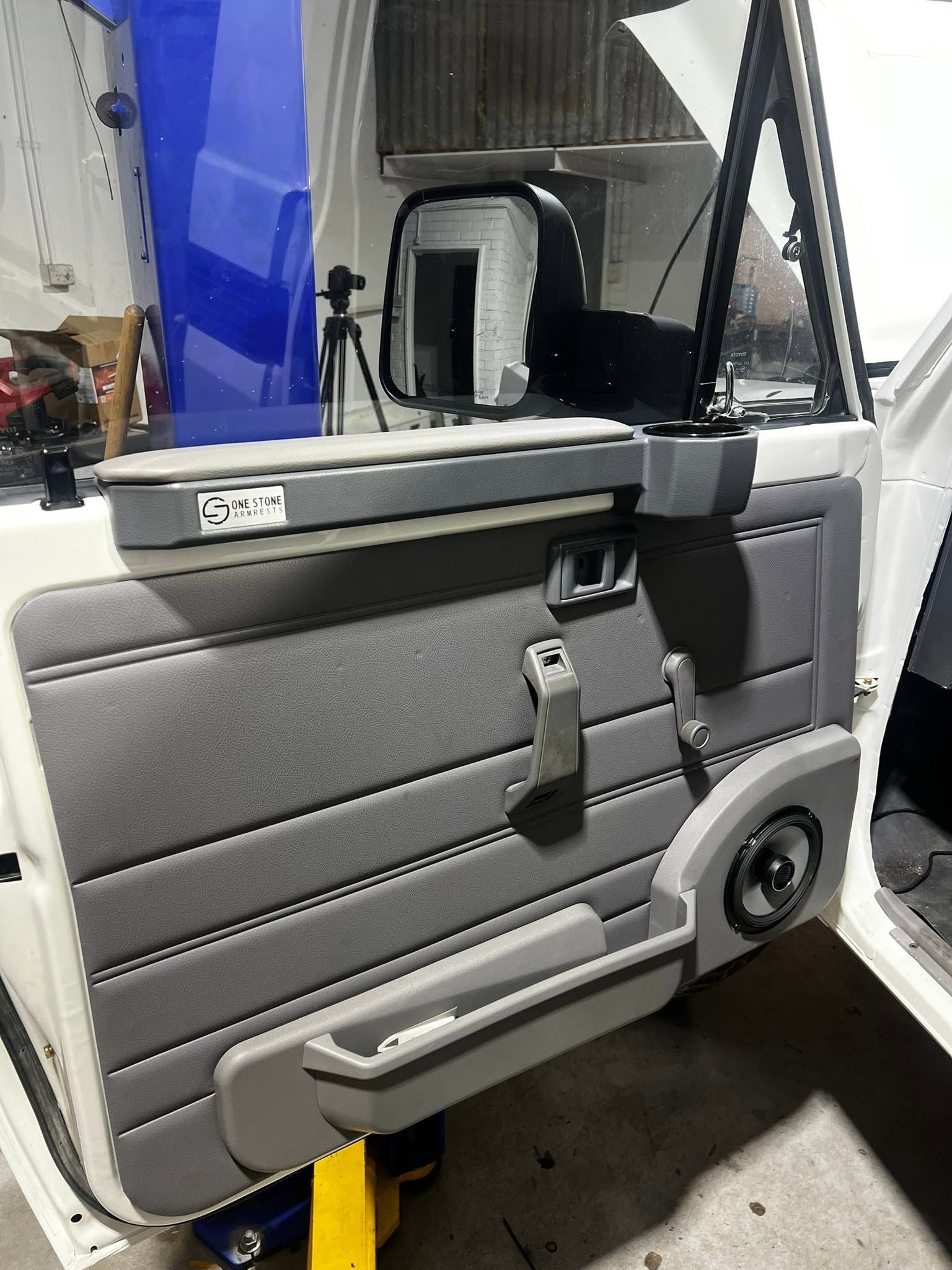 Gray car door panel with armrest, handle, window frame, and speaker, shown open in a garage.
