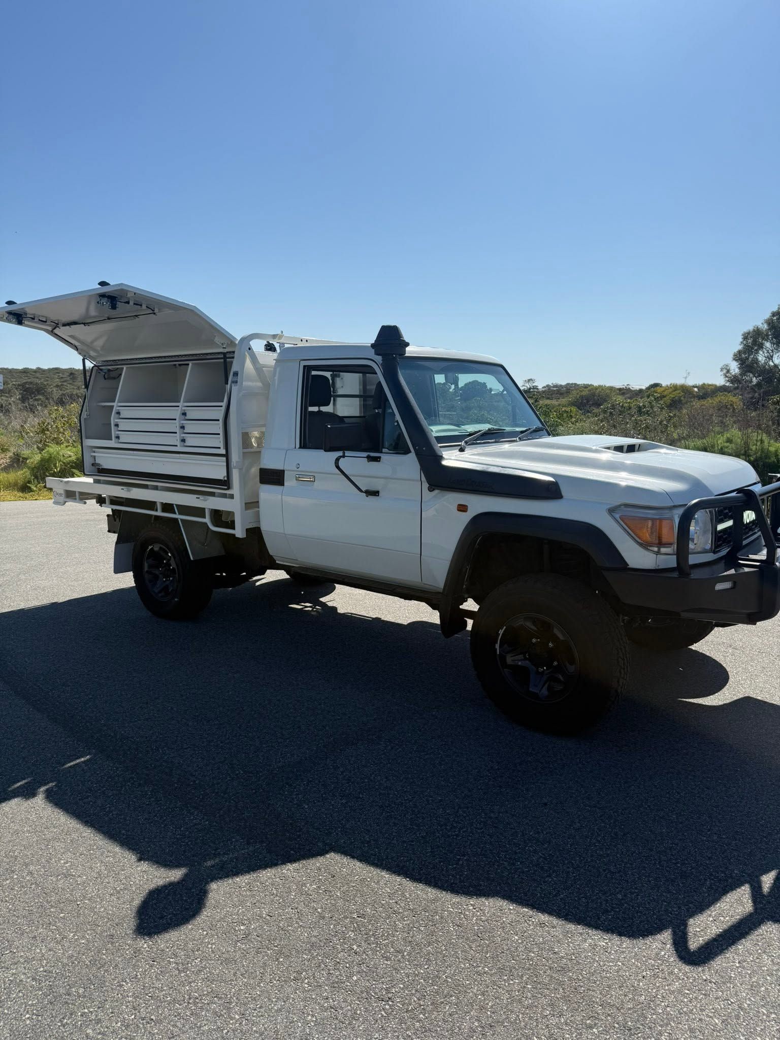 White utility truck with open side compartments parked on a sunny road under a clear blue sky.