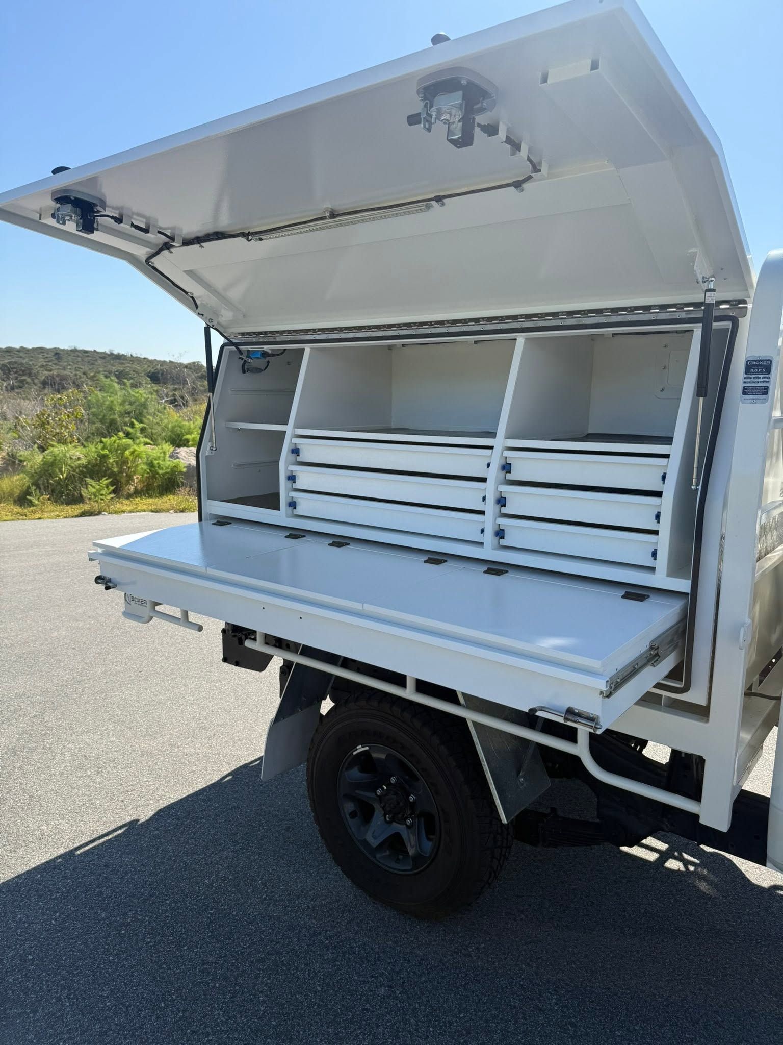Open white utility trailer with top lid raised, showing empty shelving and storage compartments outdoors.