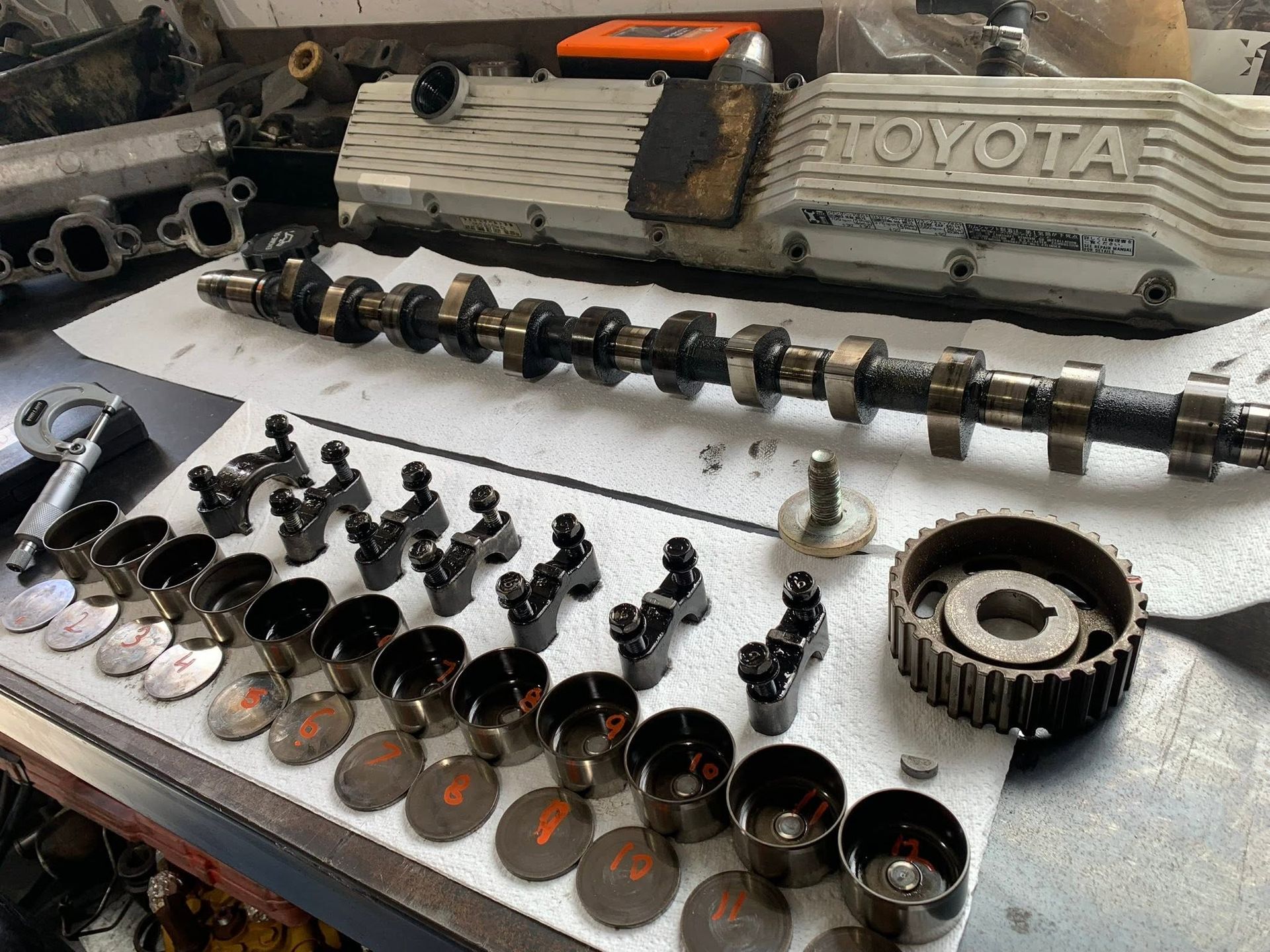 Engine parts laid out on a workbench, including camshaft, pistons, valves, and a Toyota head cover.