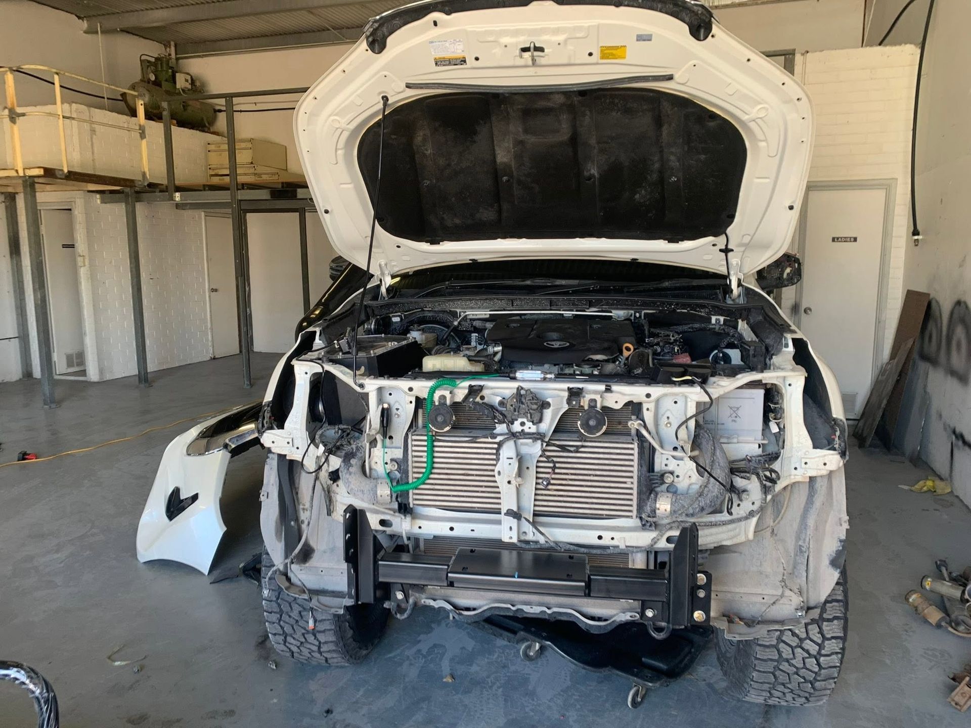 Damaged white SUV with hood open in a garage, front end stripped and exposed.