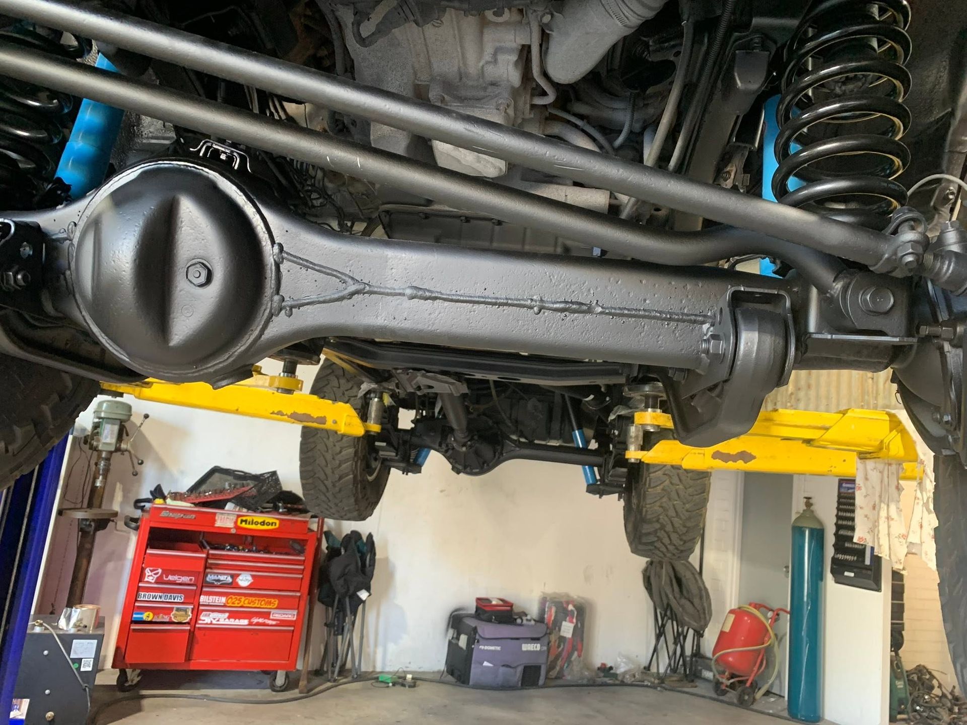 Undercarriage of a lifted vehicle in a garage, showing axle, suspension springs, and differential.