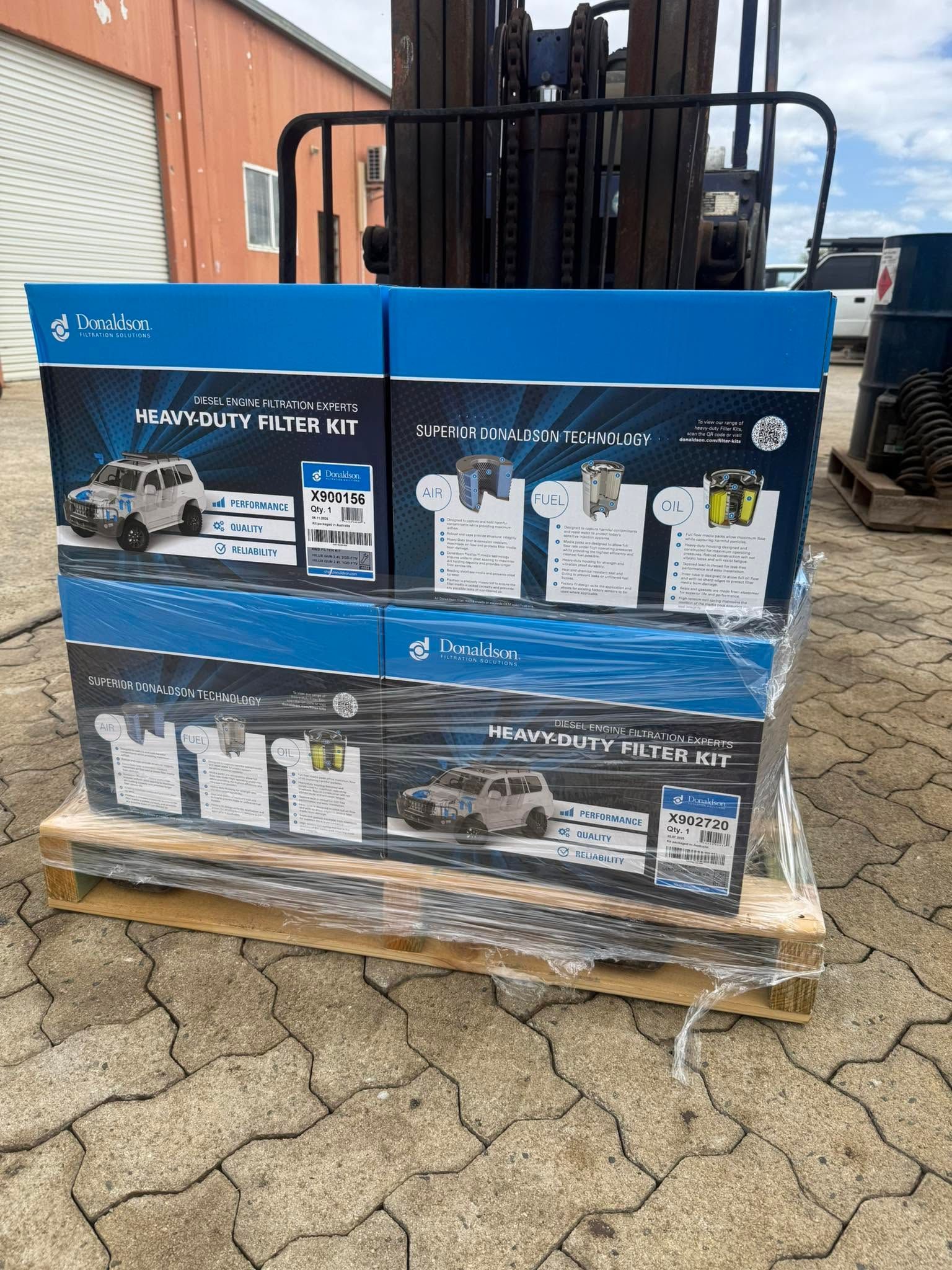 Stacked blue-and-white product boxes on a wooden pallet outdoors beside a brick building.