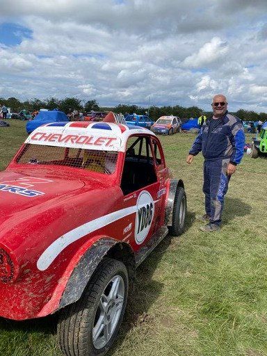 A man is standing next to a red race car in a field.