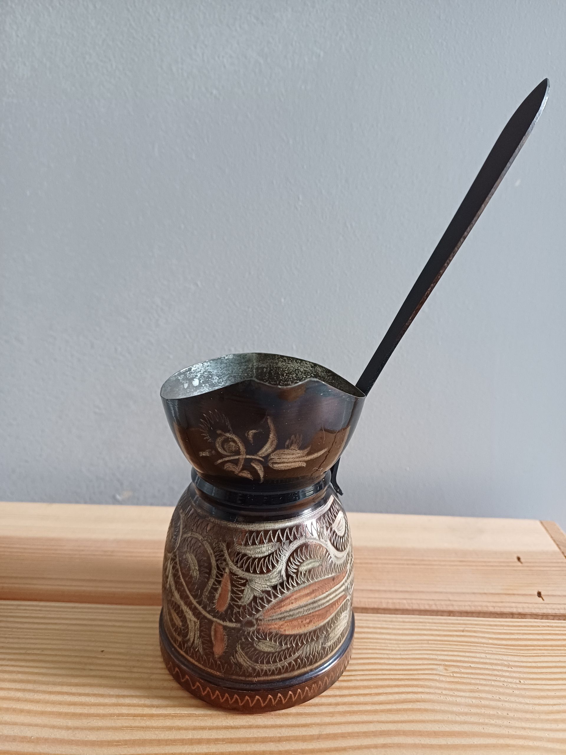 Een rijkversierde koperen cezve (koffiepot) met een lange steel, rustend op een houten ondergrond.