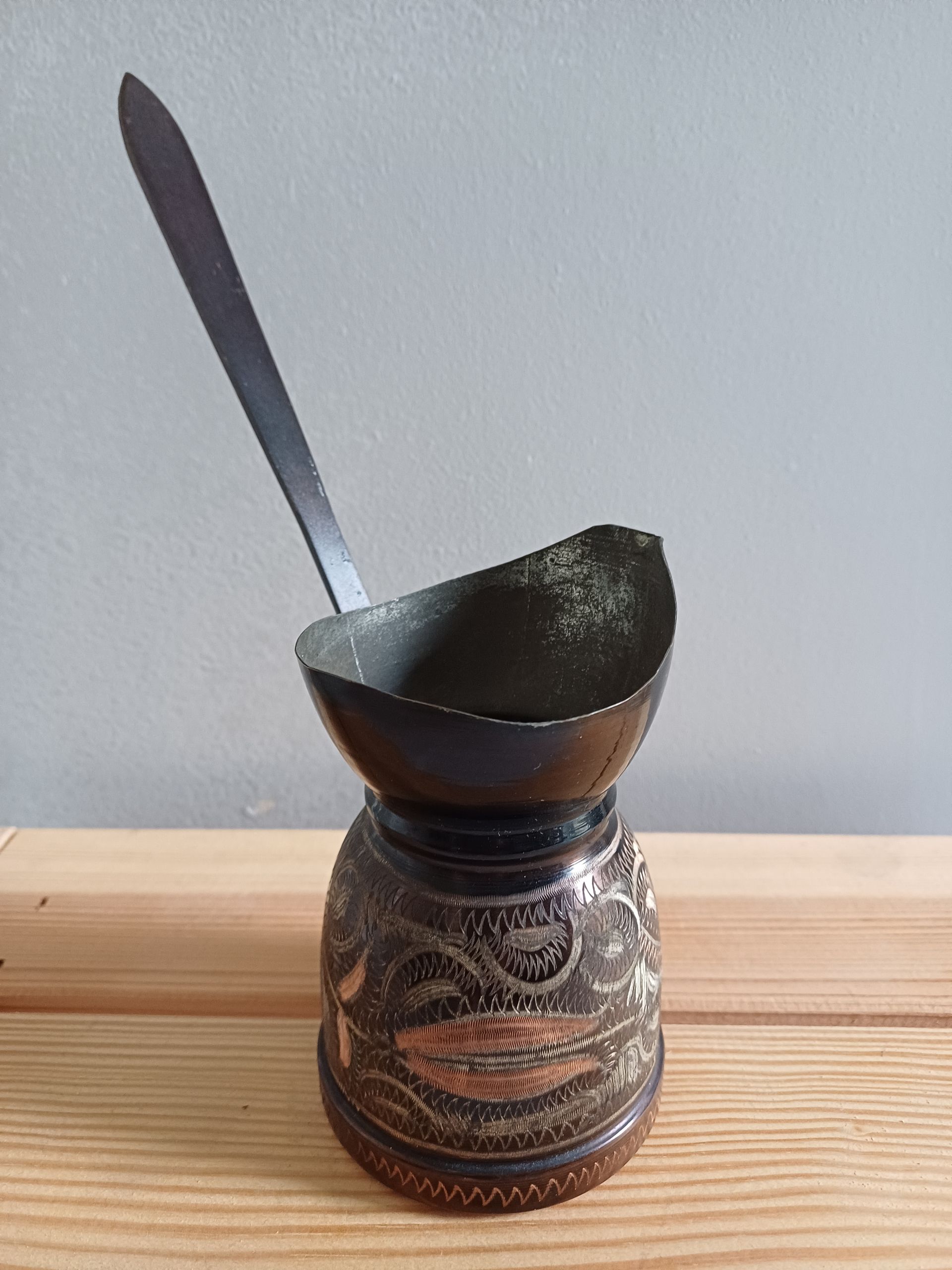 Een rijkversierde koperen cezve (koffiepot) met een lange steel, rustend op een houten ondergrond.