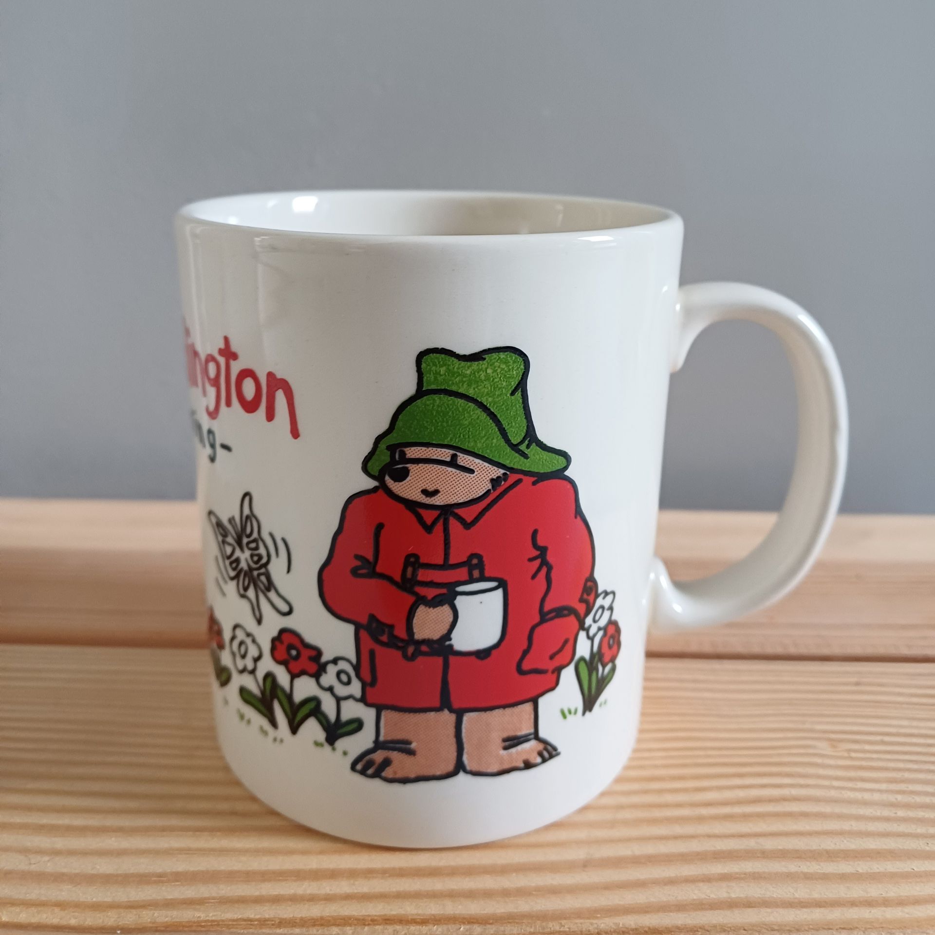 Witte mok met een afbeelding van Paddington Bear; rode jas, groene hoed, met een kopje in zijn hand.