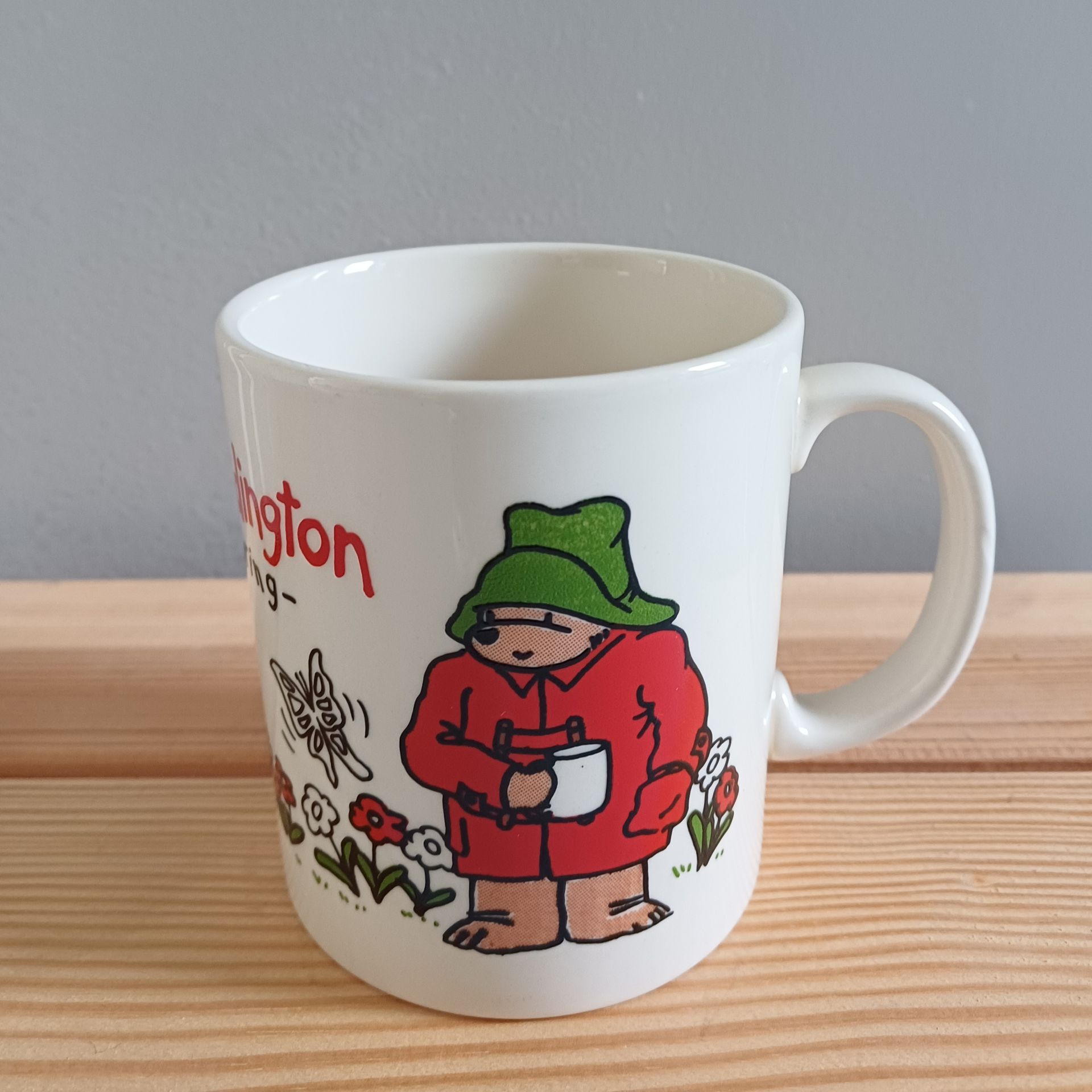 Witte mok met een afbeelding van Paddington Bear; rode jas, groene hoed, met een kopje in zijn hand.
