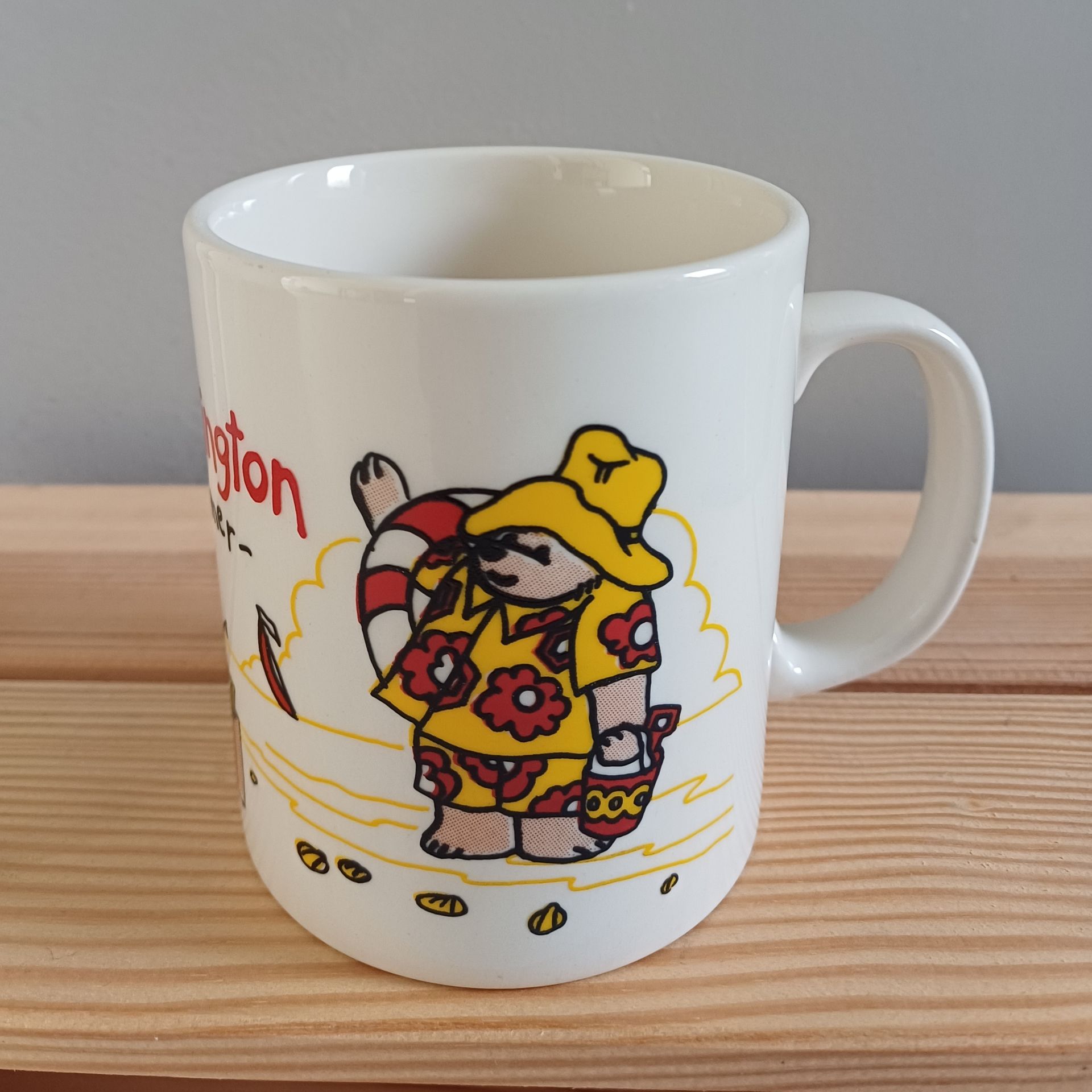 Witte mok met een cartoonbeer met een gele hoed en shirt, die een mand vasthoudt, op een strand.