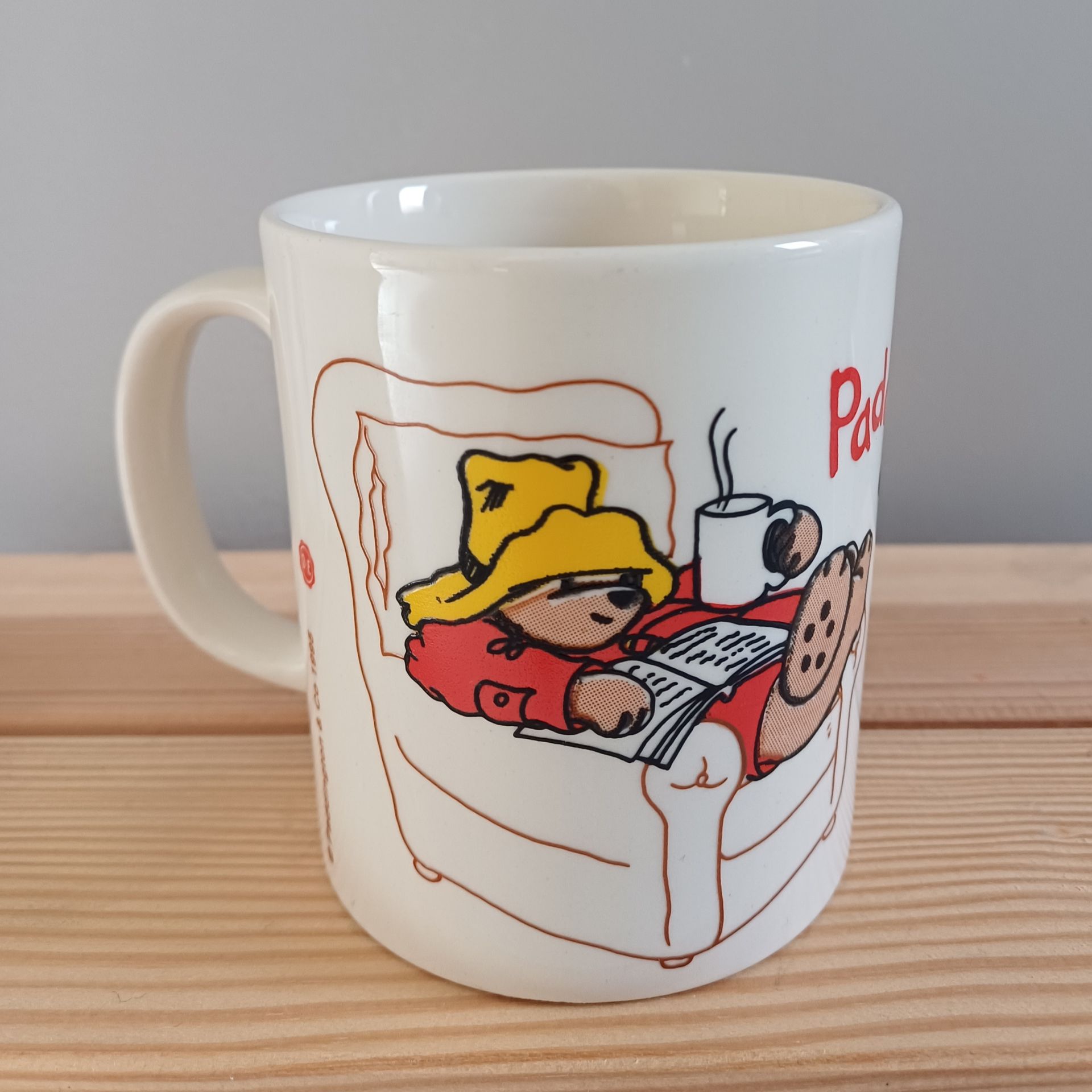 Witte mok met een cartoonbeer met een gele hoed en shirt, die een mand vasthoudt, op een strand.
