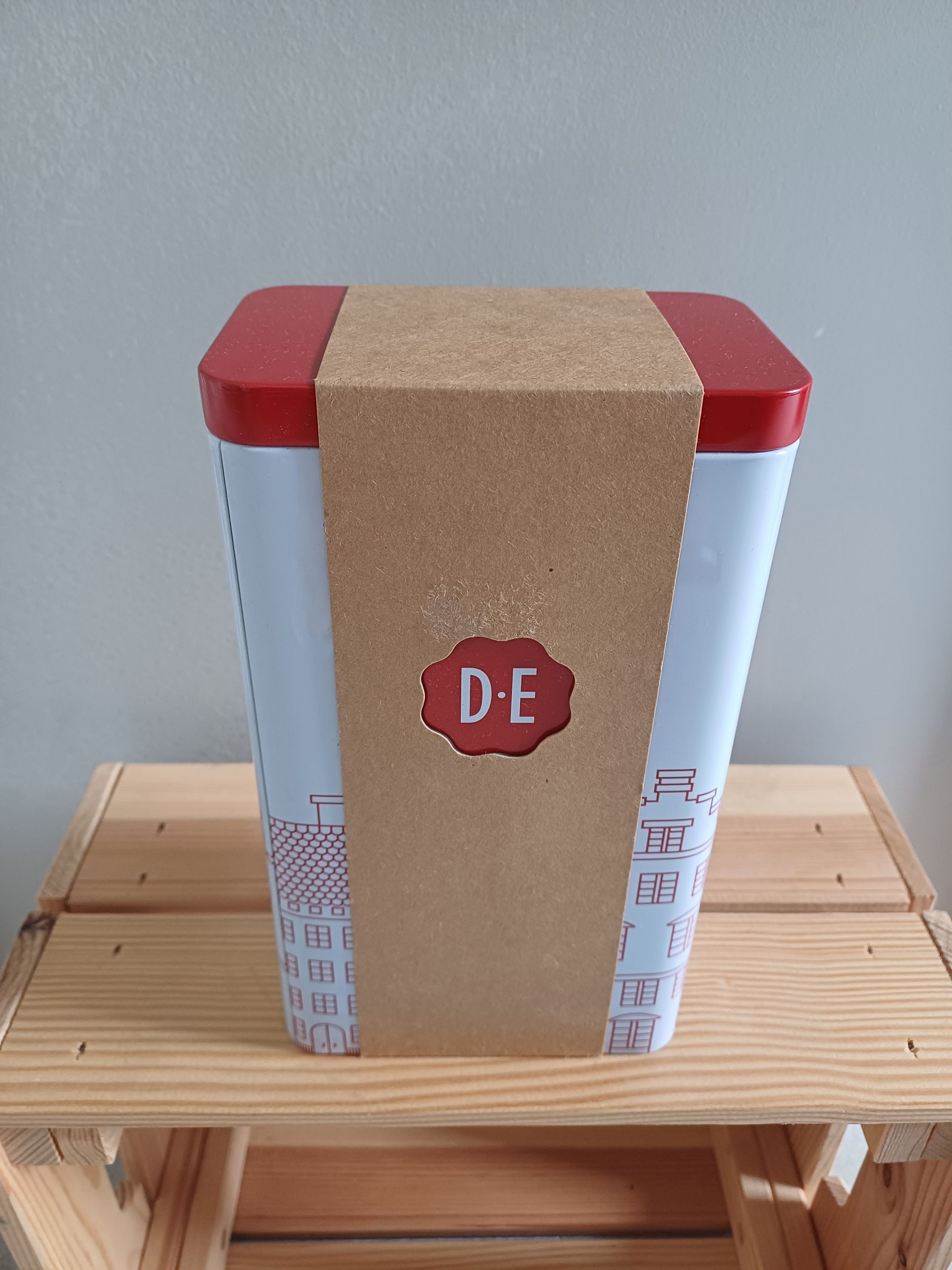 Een hoge, witte doos met een rode deksel en bodem, omwikkeld met bruin papier, staat op een houten plank.