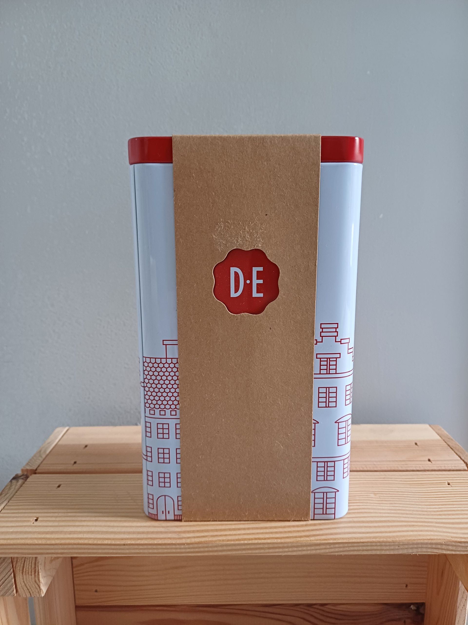 Een hoge, witte doos met een rode deksel en bodem, omwikkeld met bruin papier, staat op een houten plank.