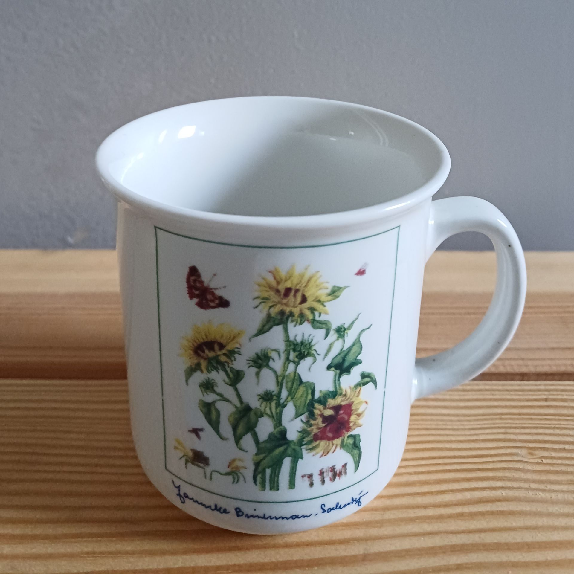 Witte mok met zonnebloemillustratie, die op een houten ondergrond staat.