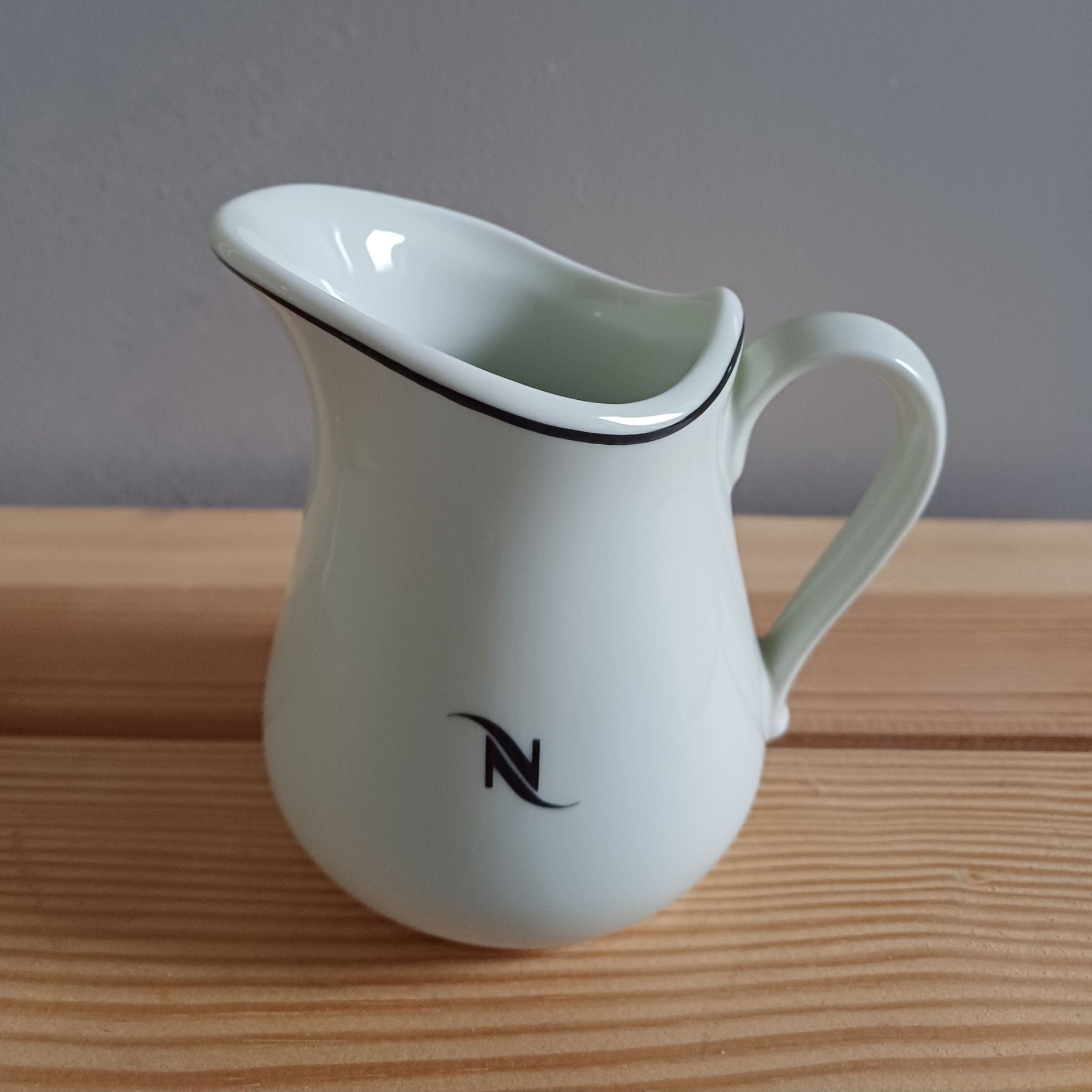 Witte Nespresso-melkkan met zwarte rand en logo, op een houten ondergrond.