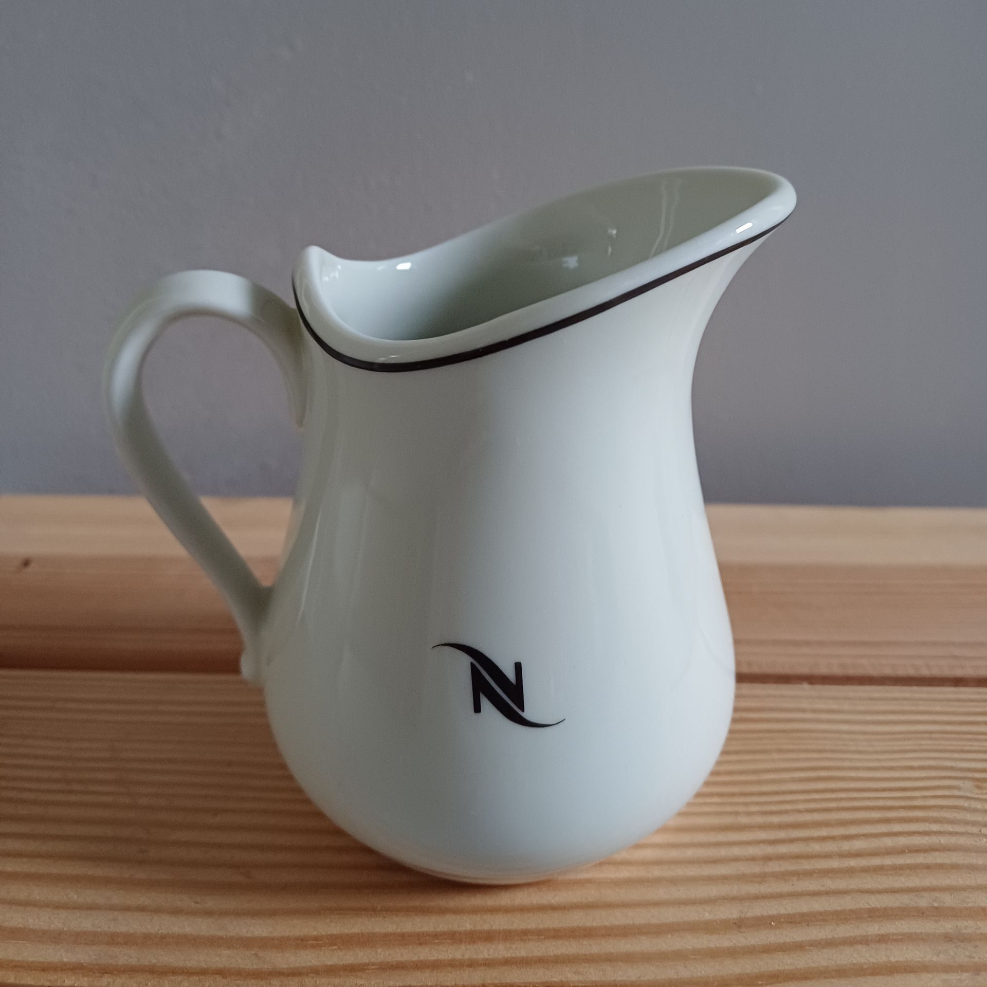 Witte Nespresso-melkkan met zwarte rand en logo, op een houten ondergrond.