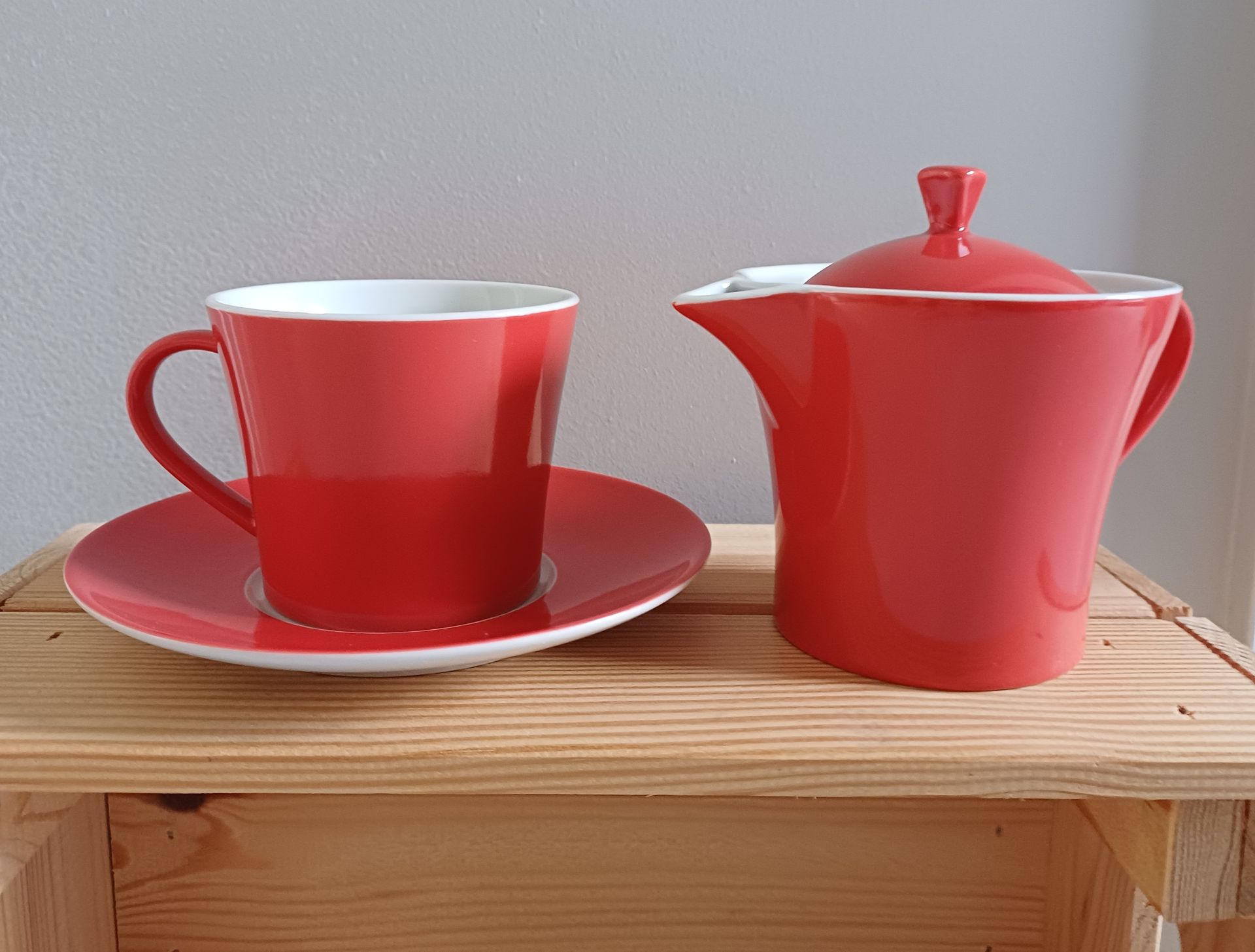 Een rood porseleinen theekopje, gestapeld op een theepot met schotel en deksel, staat op een houten ondergrond.