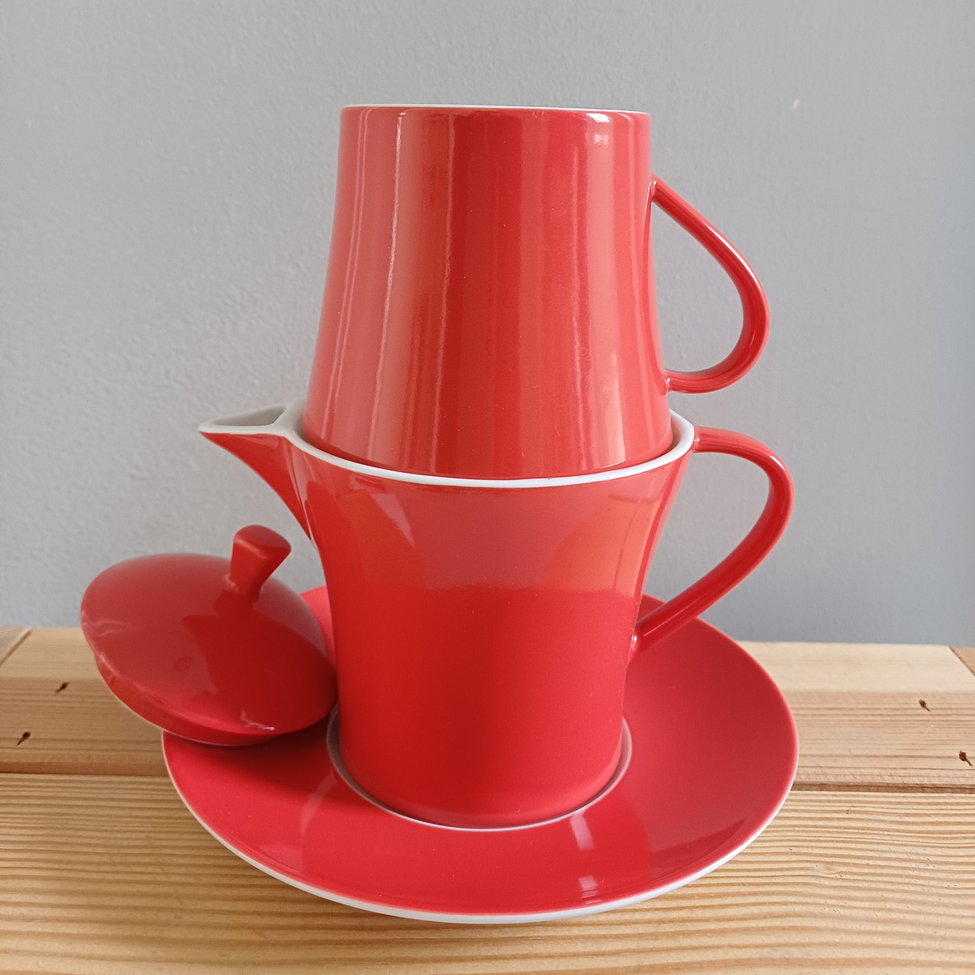 Een rood porseleinen theekopje, gestapeld op een theepot met schotel en deksel, staat op een houten ondergrond.