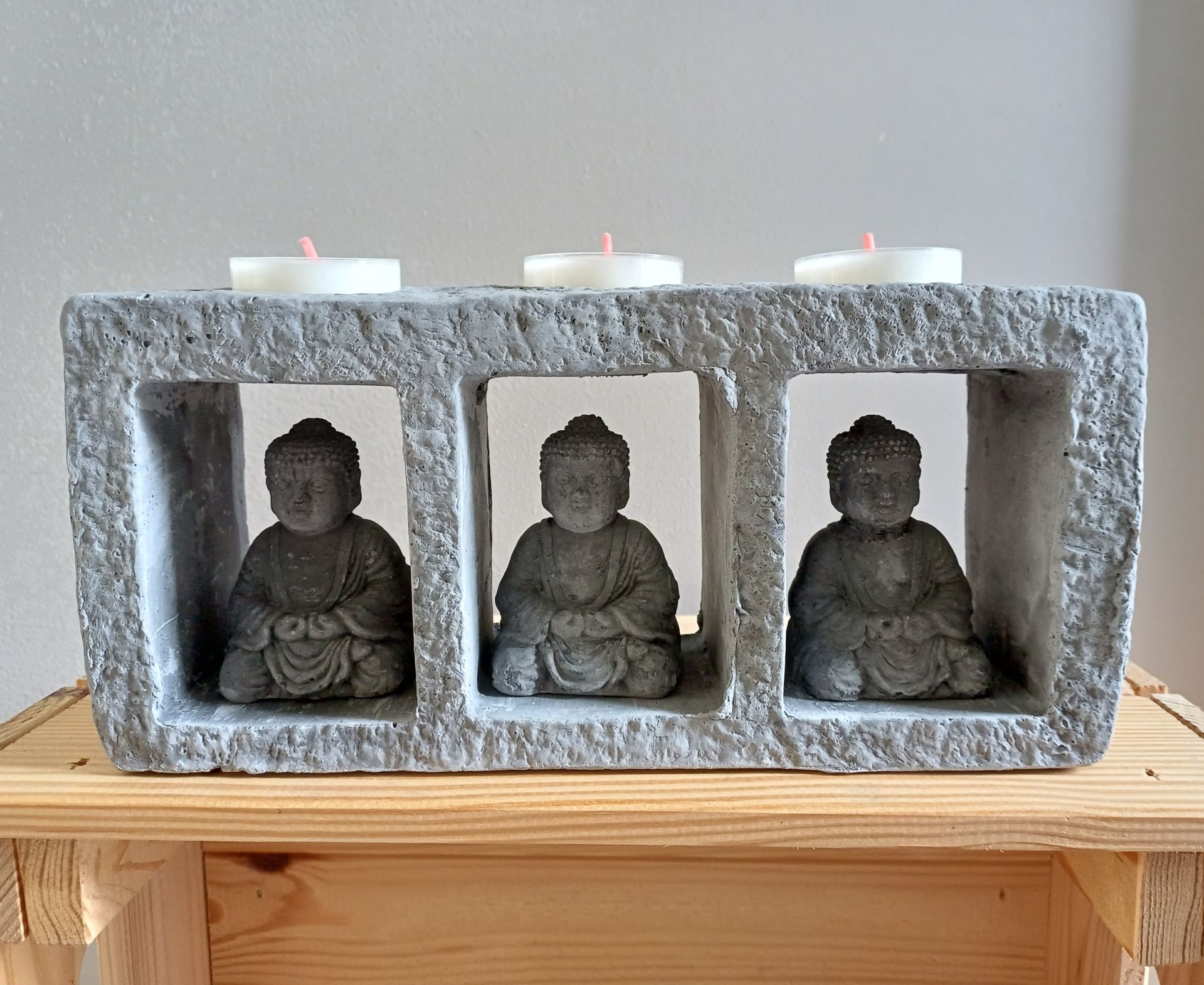 Boeddhabeeldje als kaarsenhouder: drie stenen Boeddha's in een grijs blok met kaarsen erop, op een houten plank.