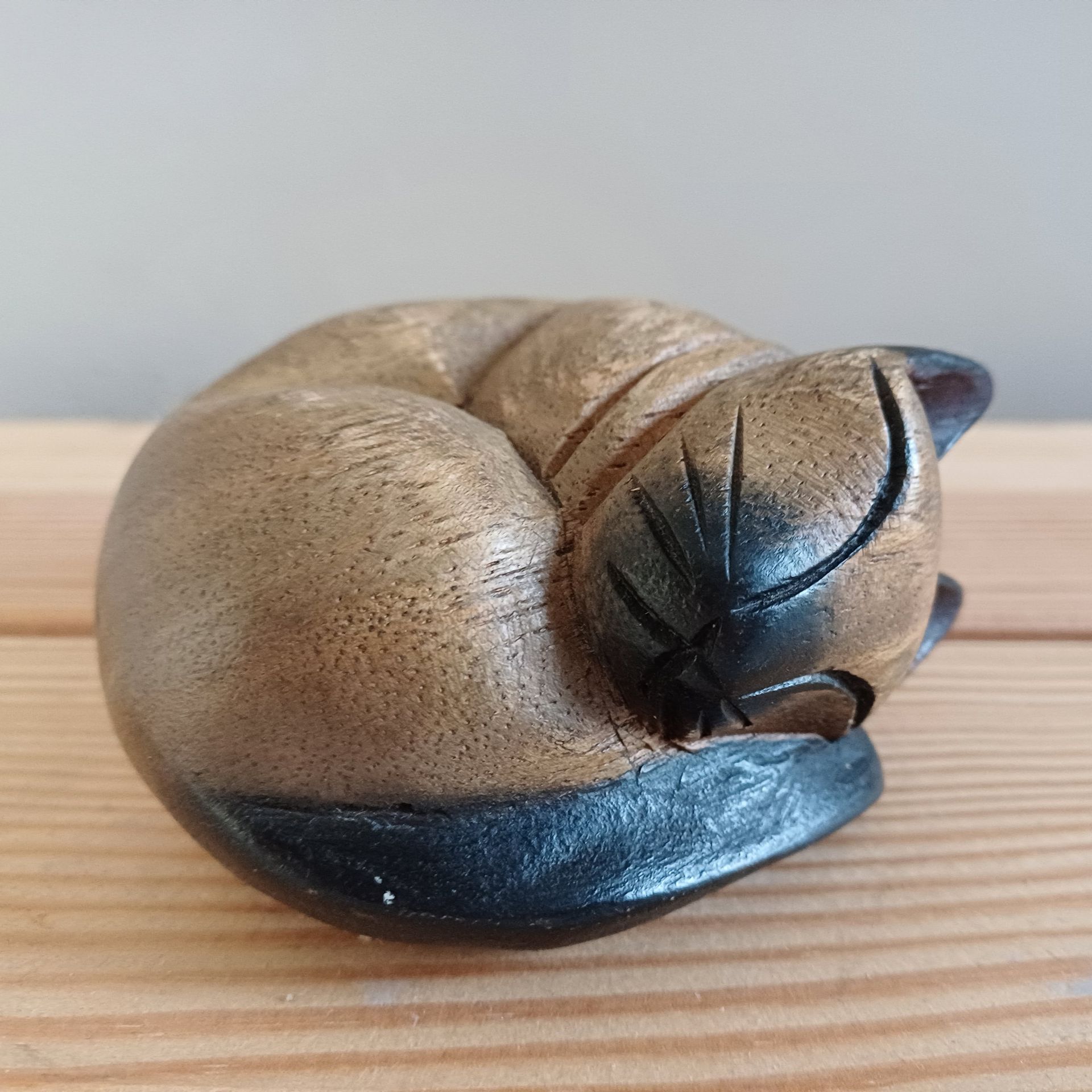 Houten beeldje van een opgerolde kat, bruin lichaam met zwarte accenten, liggend op een houten ondergrond.