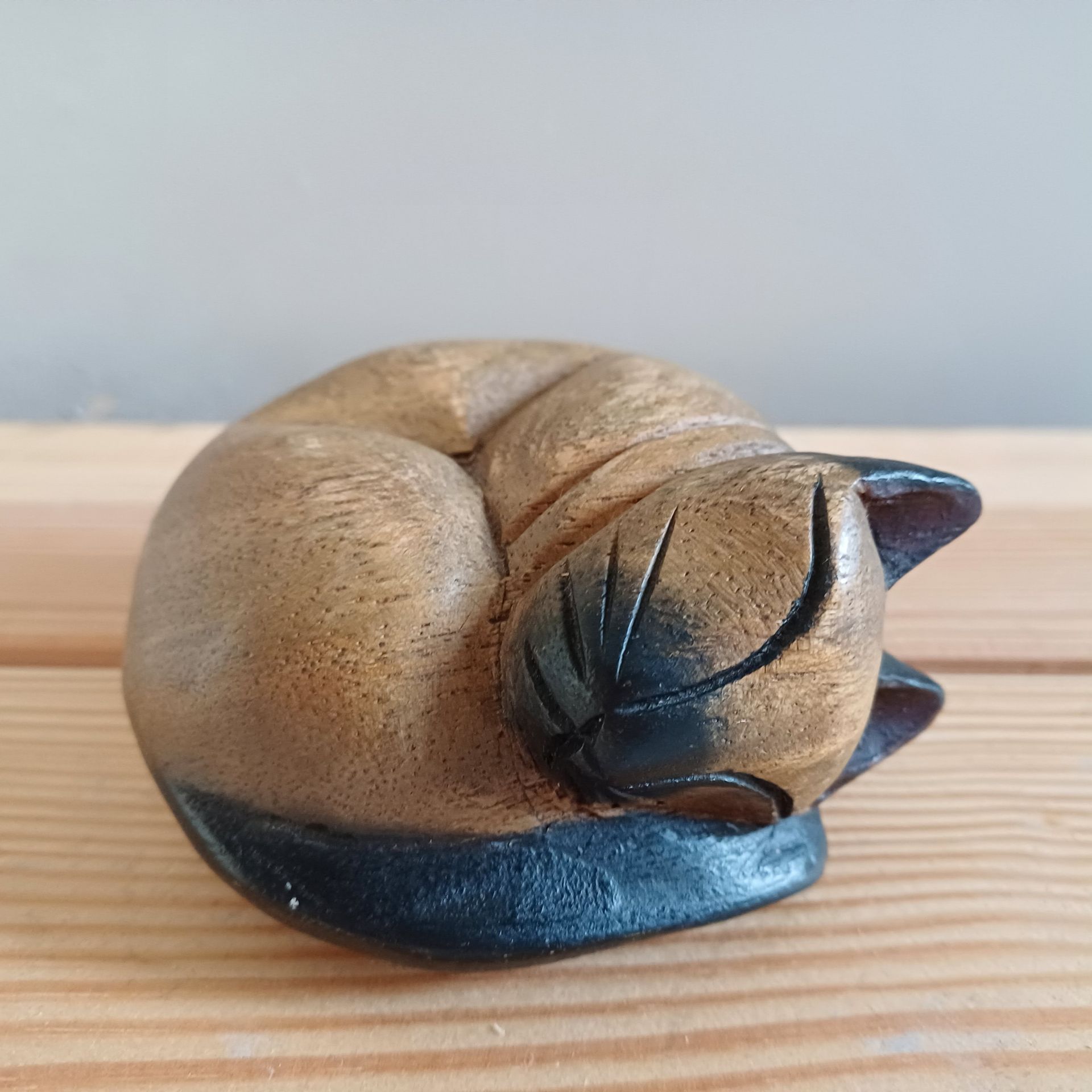 Houten beeldje van een opgerolde kat, bruin lichaam met zwarte accenten, liggend op een houten ondergrond.