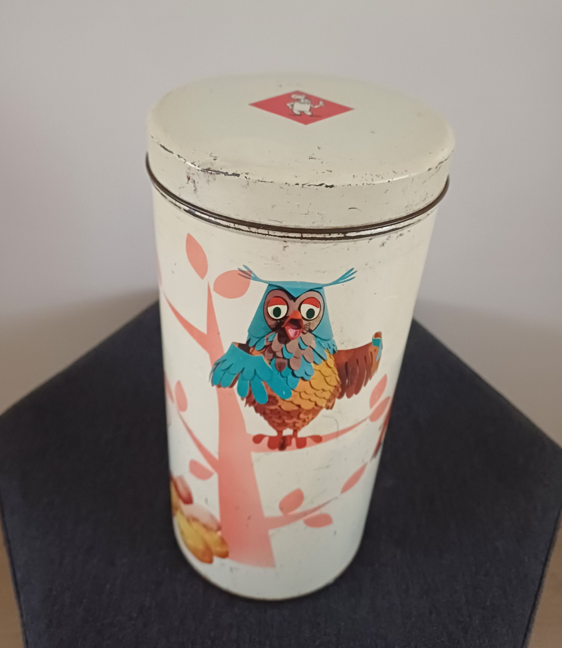 Blikje met een afbeelding van een uil die op een boomtak zit, wit, met een ronde bovenkant en een rode ruit erop.