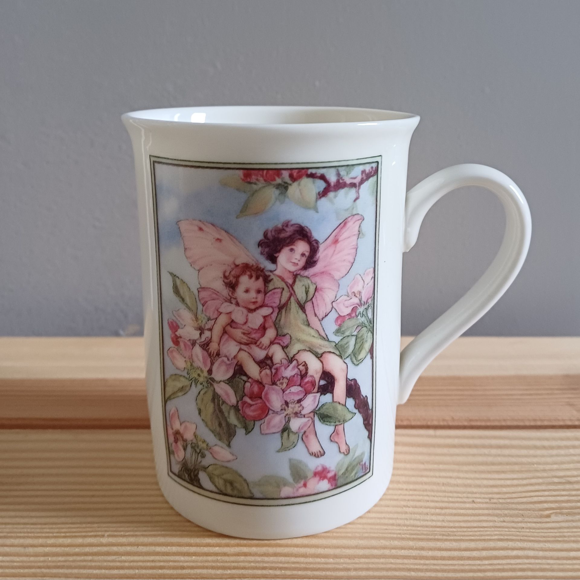 Witte mok met feeënillustratie; twee figuren met vleugels tussen roze bloemen, met een blauwe lucht op de achtergrond.