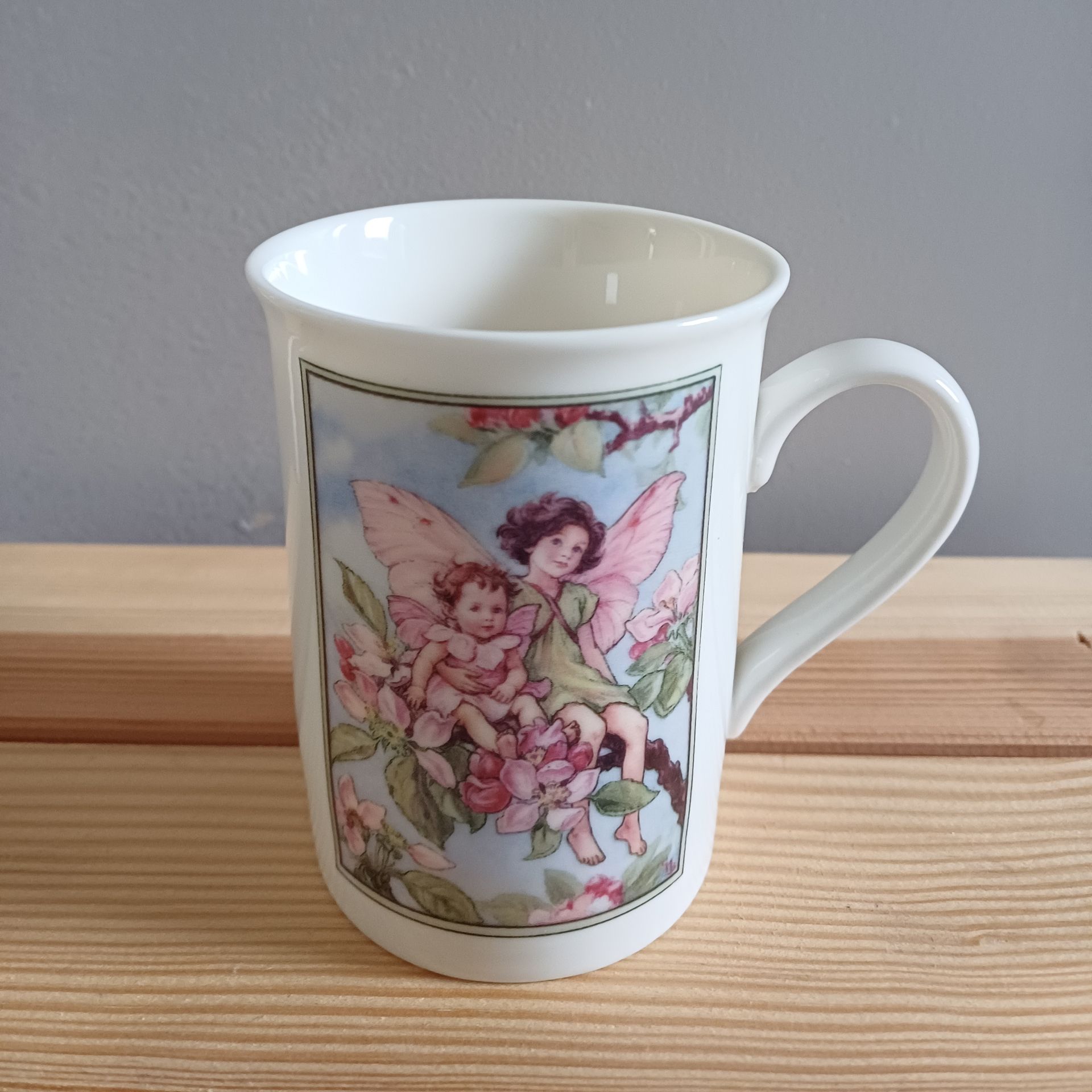 Witte mok met feeënillustratie; twee figuren met vleugels tussen roze bloemen, met een blauwe lucht op de achtergrond.