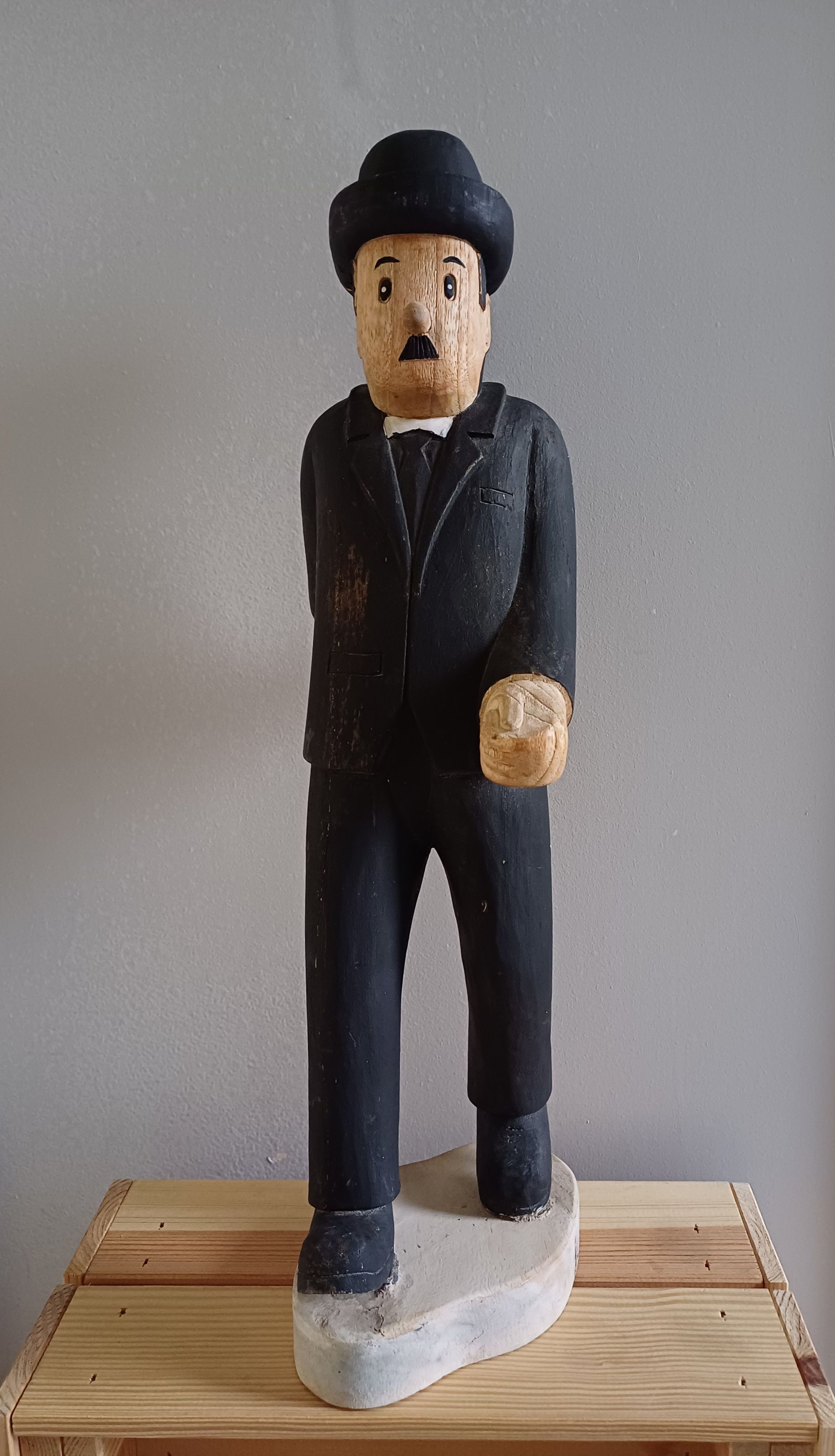 Houten beeldhouwwerk van Charlie Chaplin in een zwart pak met bolhoed, op een stenen sokkel.