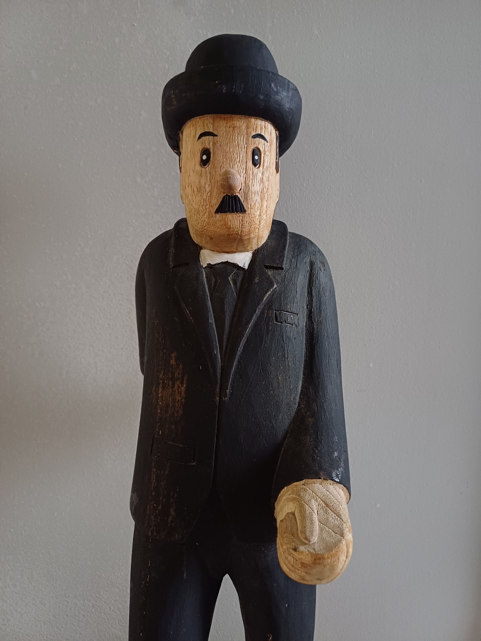 Houten beeldhouwwerk van Charlie Chaplin in een zwart pak met bolhoed, op een stenen sokkel.