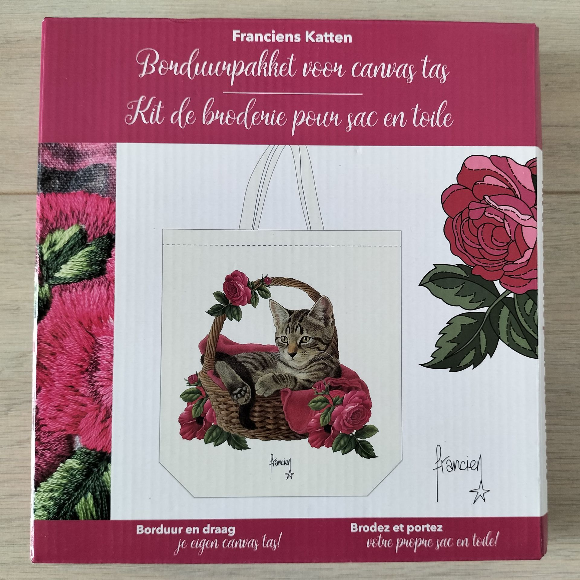 Een borduurpakket in een doos met een kat in een mandje, omringd door rozen, op een draagtas. Roze en wit.